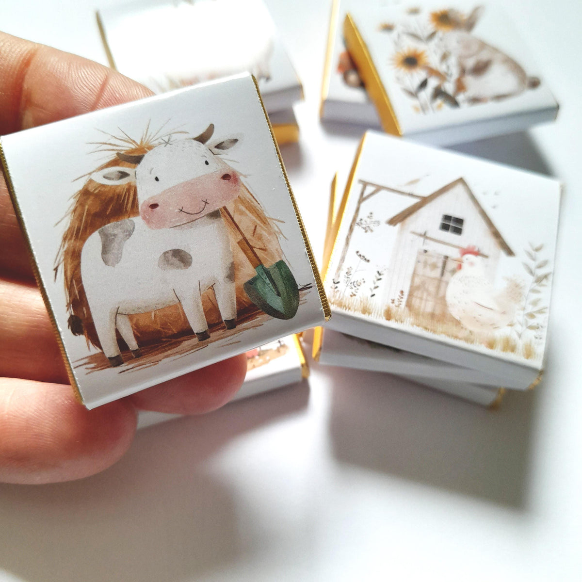 May include: A stack of small white boxes with gold trim, each featuring a different farm animal illustration. The top box shows a white cow with brown spots holding a green shovel, standing in a field of brown hay.
