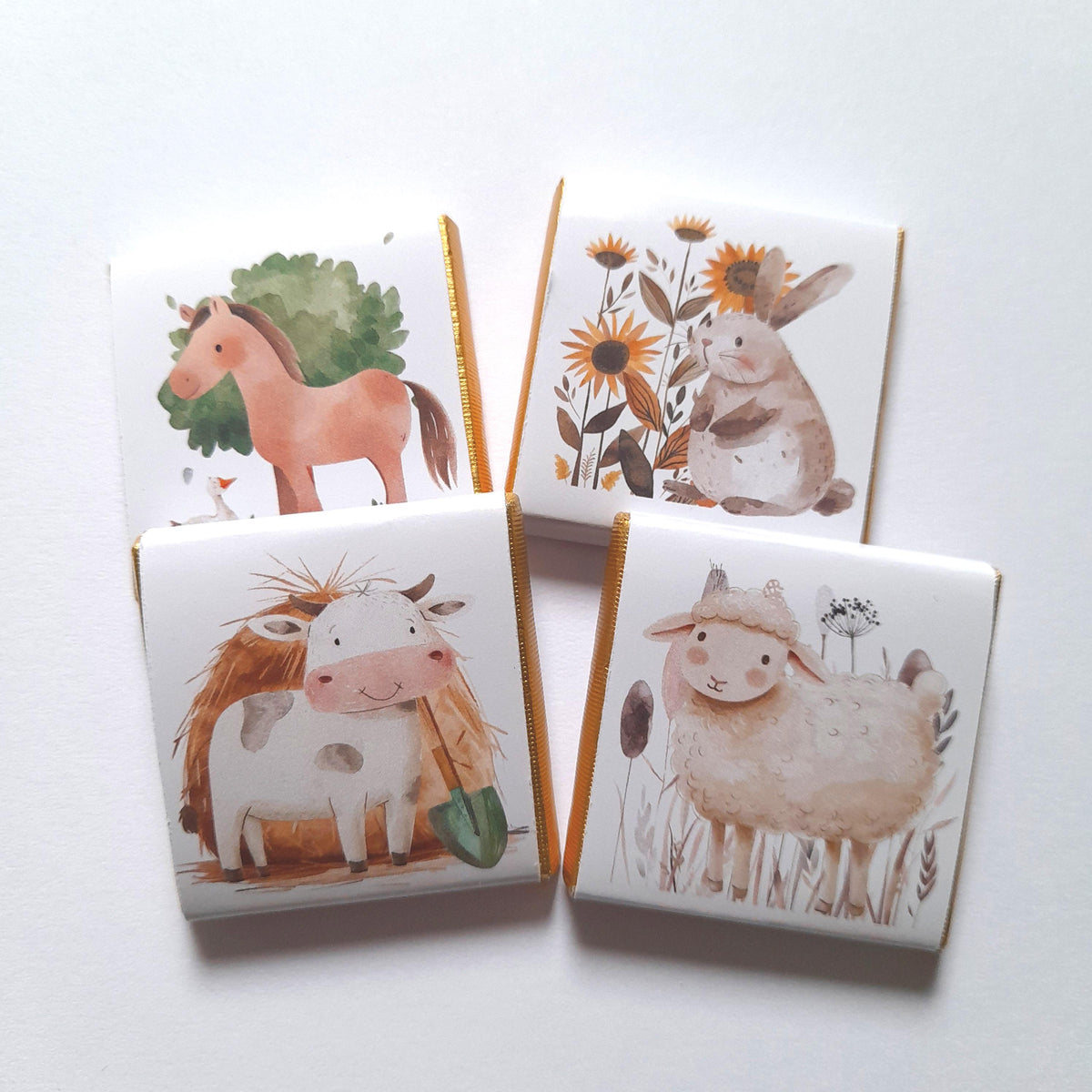 May include: Four small white chocolate bars with gold foil trim. Each bar features a different farm animal illustration: a horse, a cow, a sheep, and a rabbit. The rabbit is surrounded by flowers.