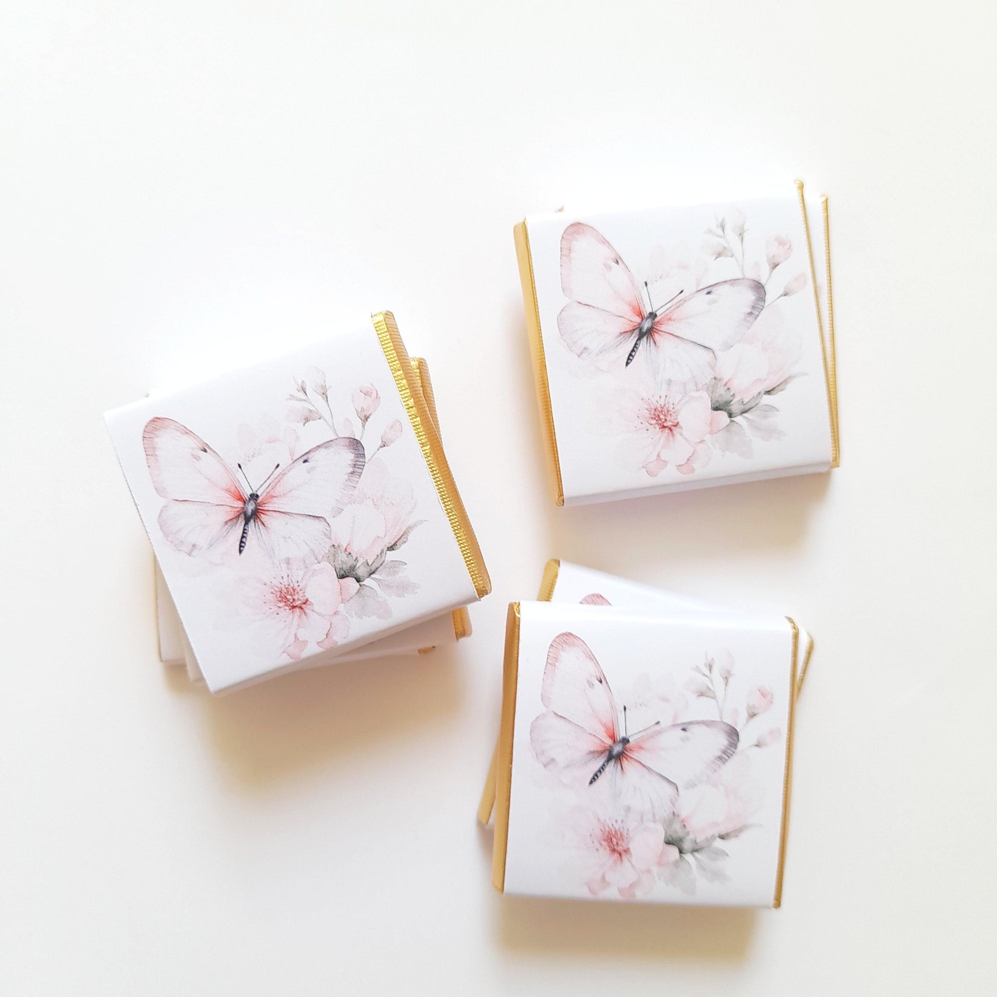 May include: A stack of white chocolate bar wrappers with a gold trim. Each wrapper features a watercolour illustration of a butterfly with pink flowers.