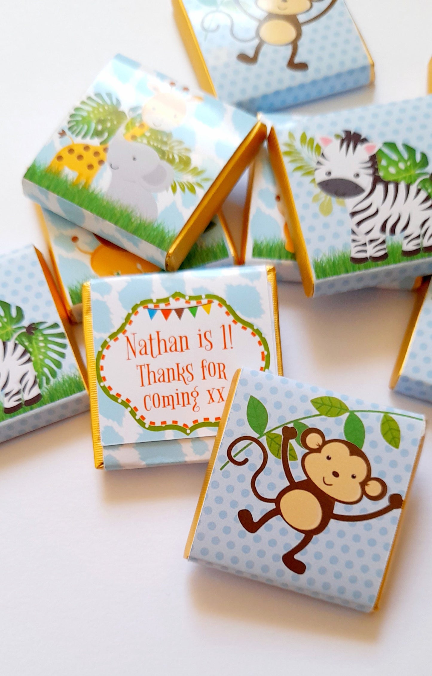May include: A stack of blue and white chocolate bar wrappers with a jungle animal theme. The wrappers feature a monkey, zebra, giraffe, and elephant. One wrapper has a personalised message that says "Nathan is 1! Thanks for coming xx".