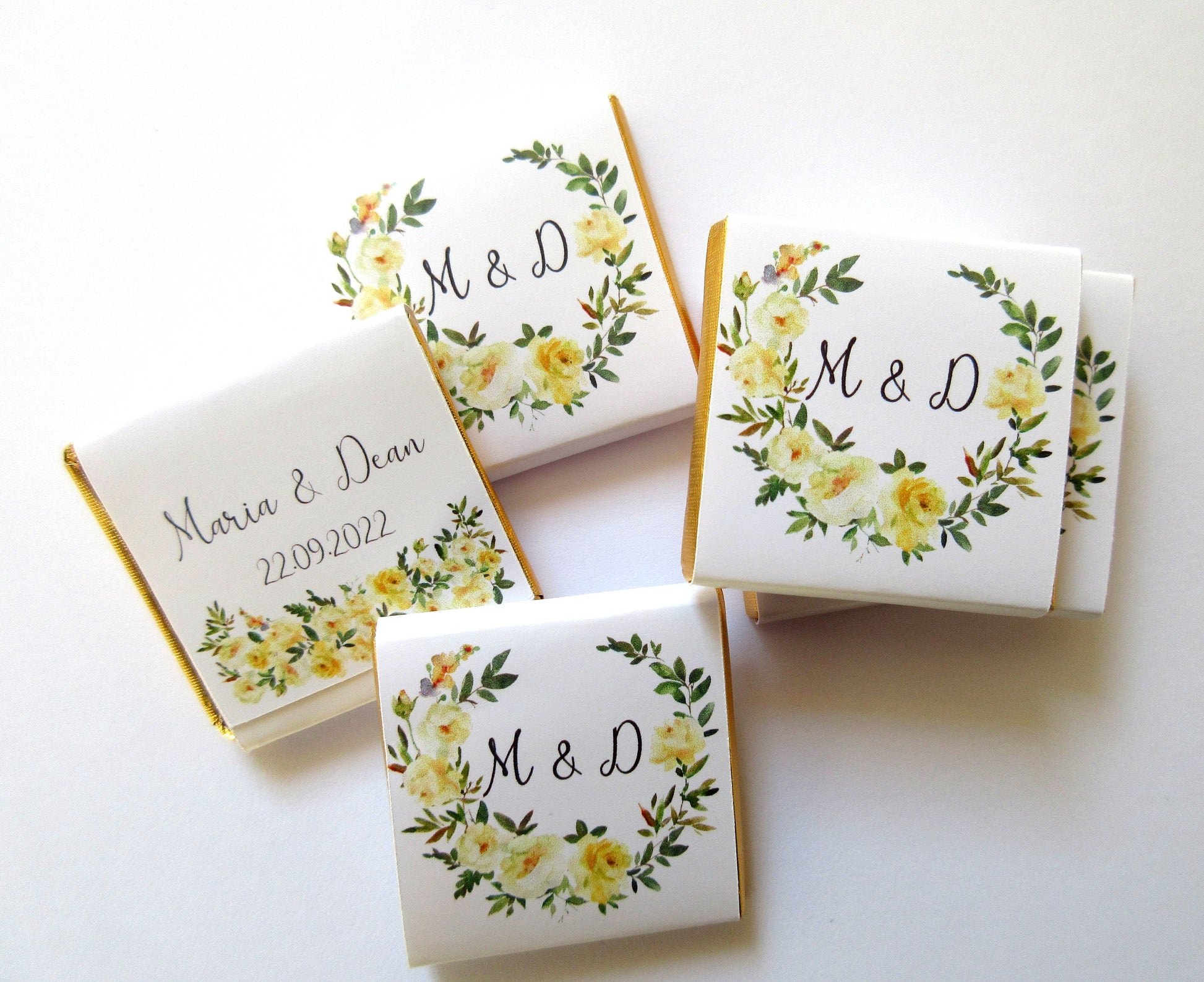 May include: A set of five small white chocolate bars with gold foil wrappers. The wrappers feature a floral design with yellow roses and green leaves. The text "Maria & Dean 22.09.2022" and "M & D" are printed on the wrappers.