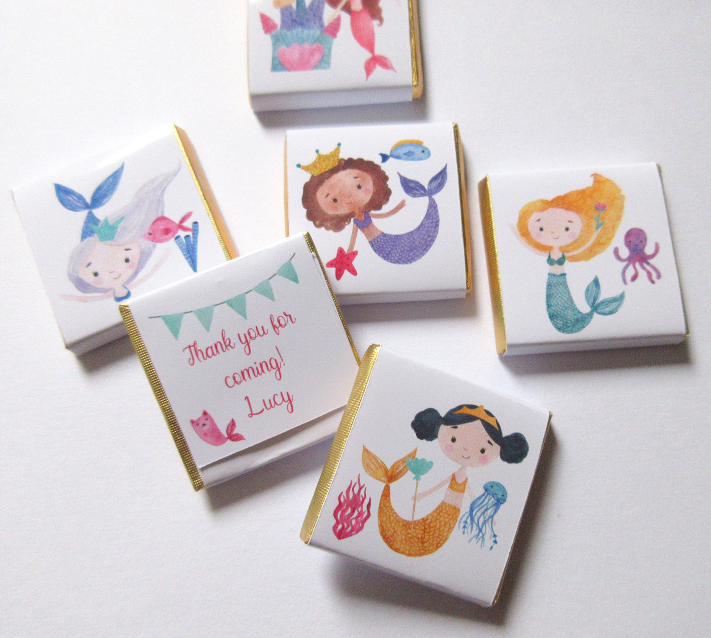May include: Five small white chocolate bars with gold foil wrappers. Each bar features a different illustration of a mermaid with a crown, holding a starfish, a flower, or a jellyfish. The text "Thank you for coming! Lucy" is printed on one of the bars.
