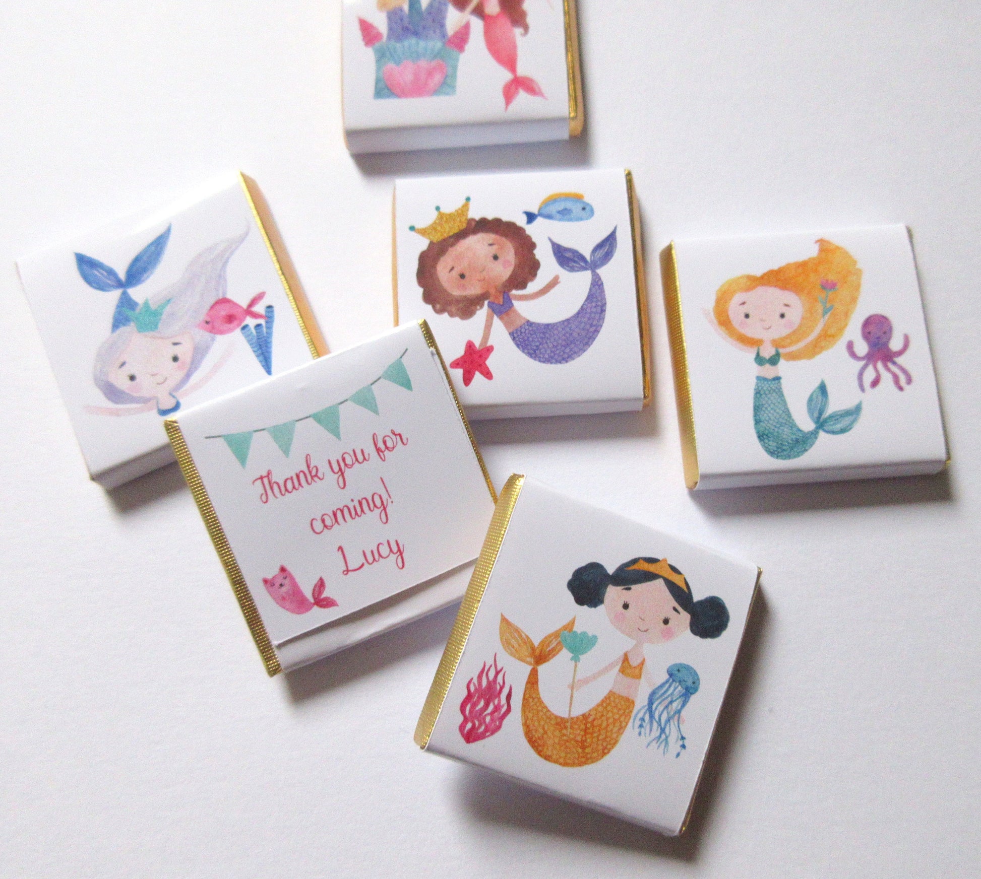 May include: Five small white chocolate bars with gold foil wrappers. Each bar features a different illustration of a mermaid with a crown, holding a starfish, a flower, or a jellyfish. The text "Thank you for coming! Lucy" is printed on one of the bars.