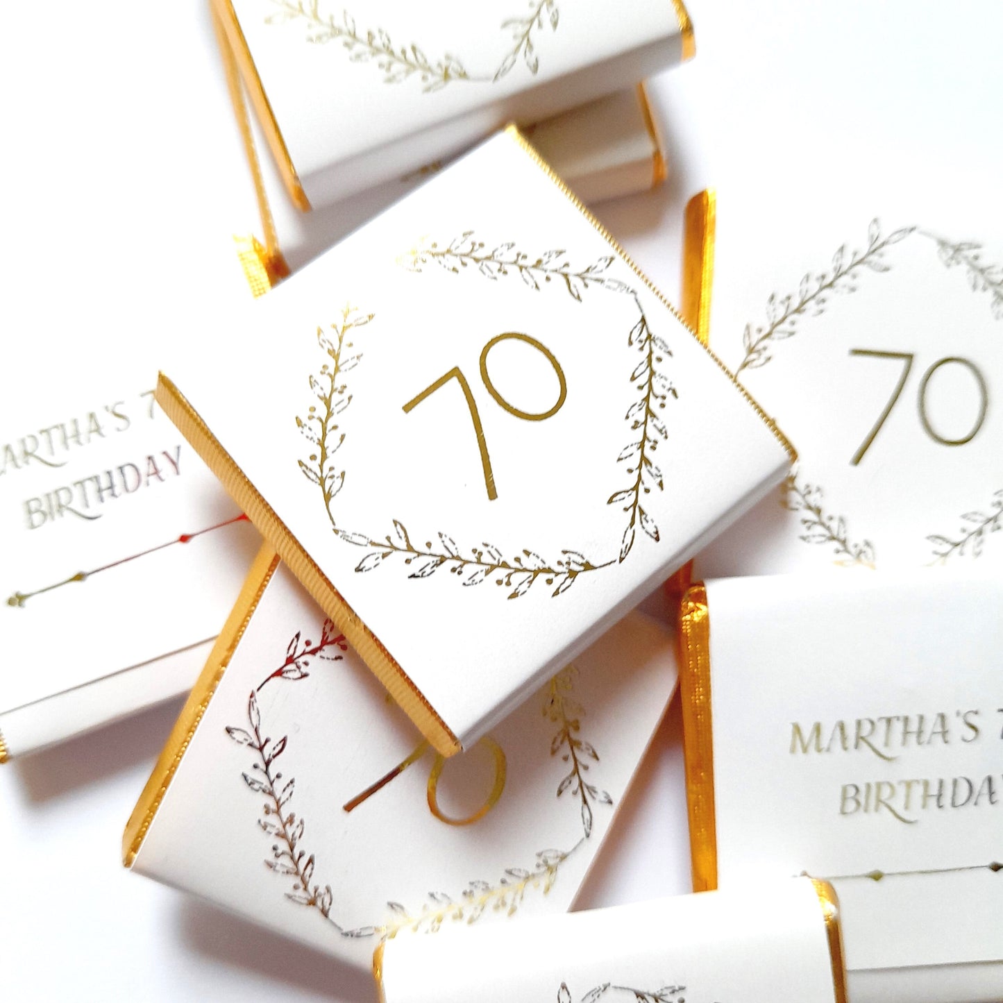 May include: A stack of white chocolate bars with gold foil wrappers. The bars are decorated with a gold number 70 inside a gold leaf wreath. The text "Martha's 70th Birthday" is printed on one of the bars.