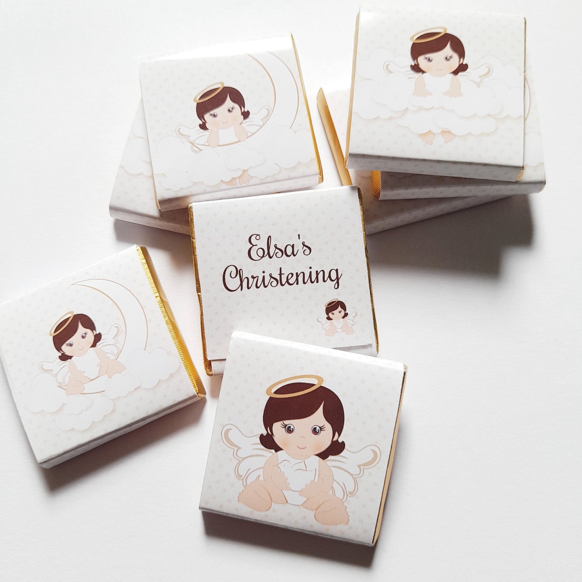 May include: White chocolate favours with gold edging, decorated with an angel illustration. Some have the text "Elsa's Christening". The angel has brown hair, a gold halo, and white wings. The background has a subtle polka dot pattern.
