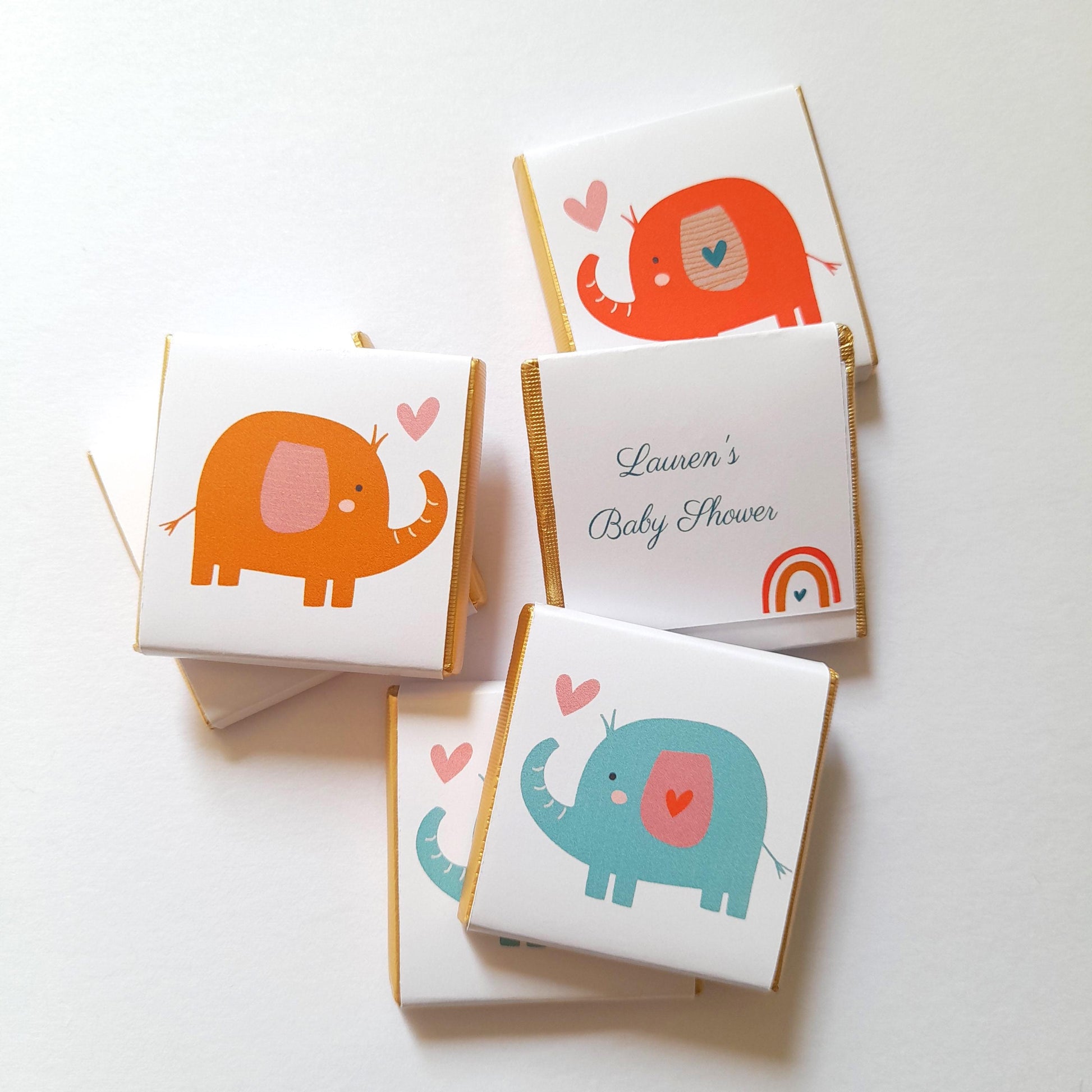 May include: A set of four small, square, white chocolate bars with gold foil wrappers. Each bar features a colourful cartoon elephant with a pink heart. One bar has the text "Lauren's Baby Shower" and a rainbow.