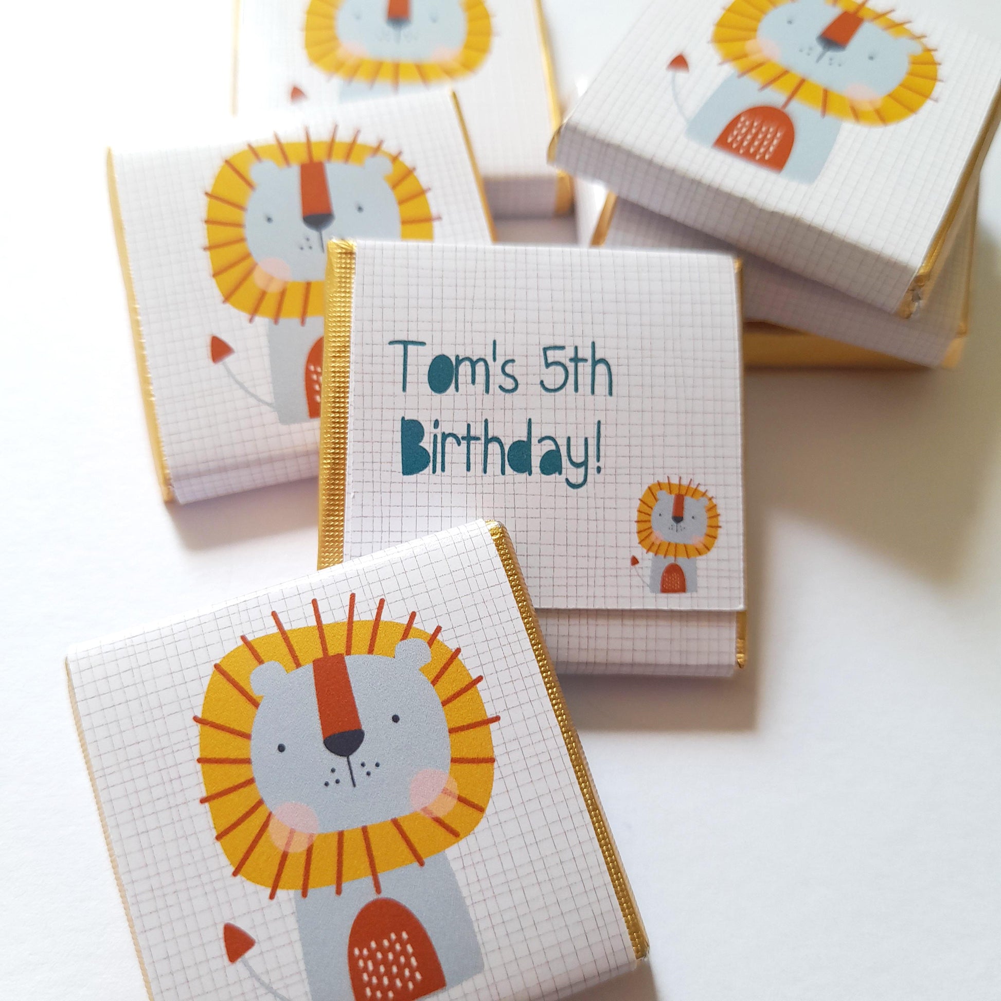 May include: White chocolate bar wrappers with a gold border and a cartoon lion design. The wrappers have the text "Tom's 5th Birthday!" printed on them.