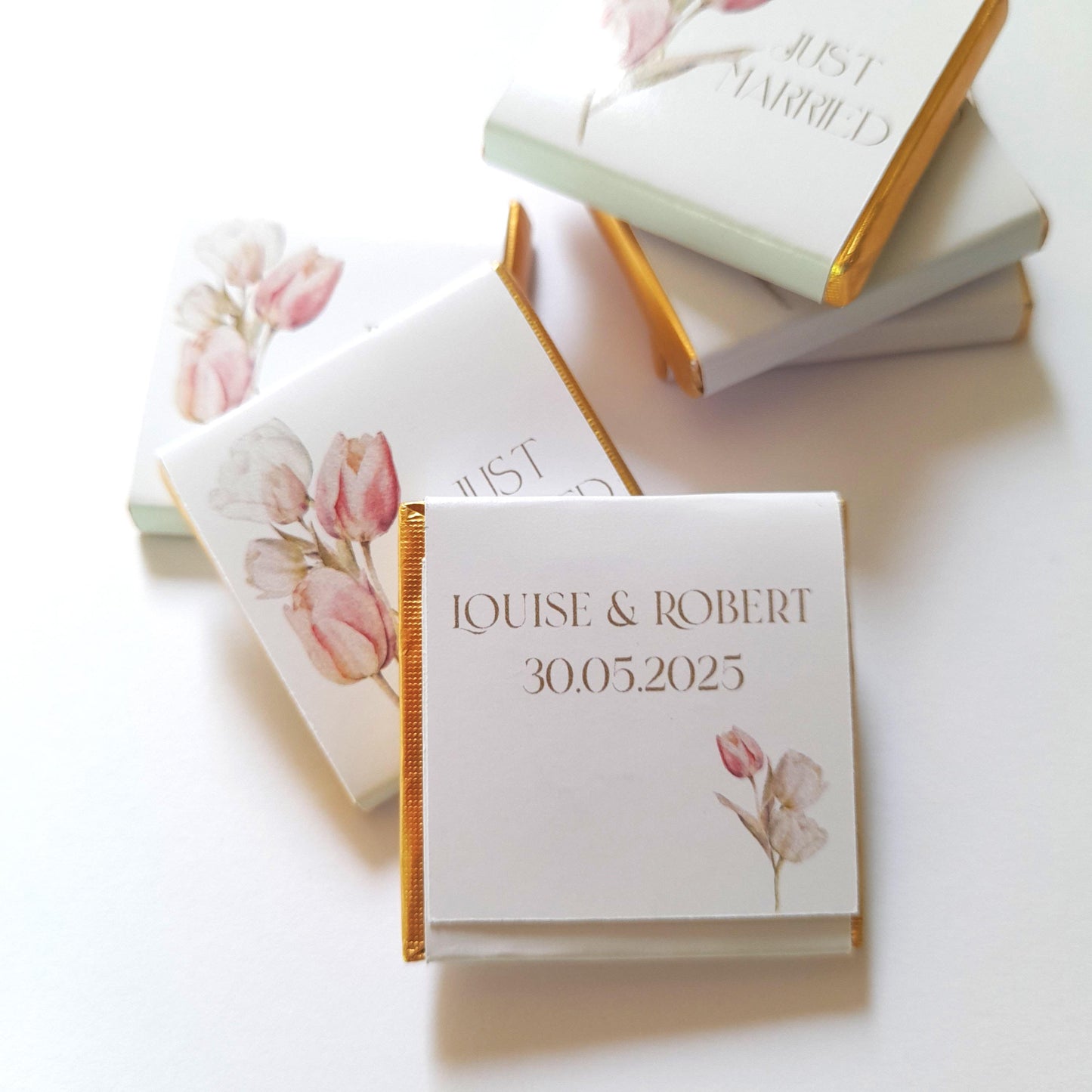 May include: White chocolate bar wrapped in white paper with gold trim. The paper features a watercolour floral design with pink and white flowers and the text "Louise & Robert 30.05.2025".