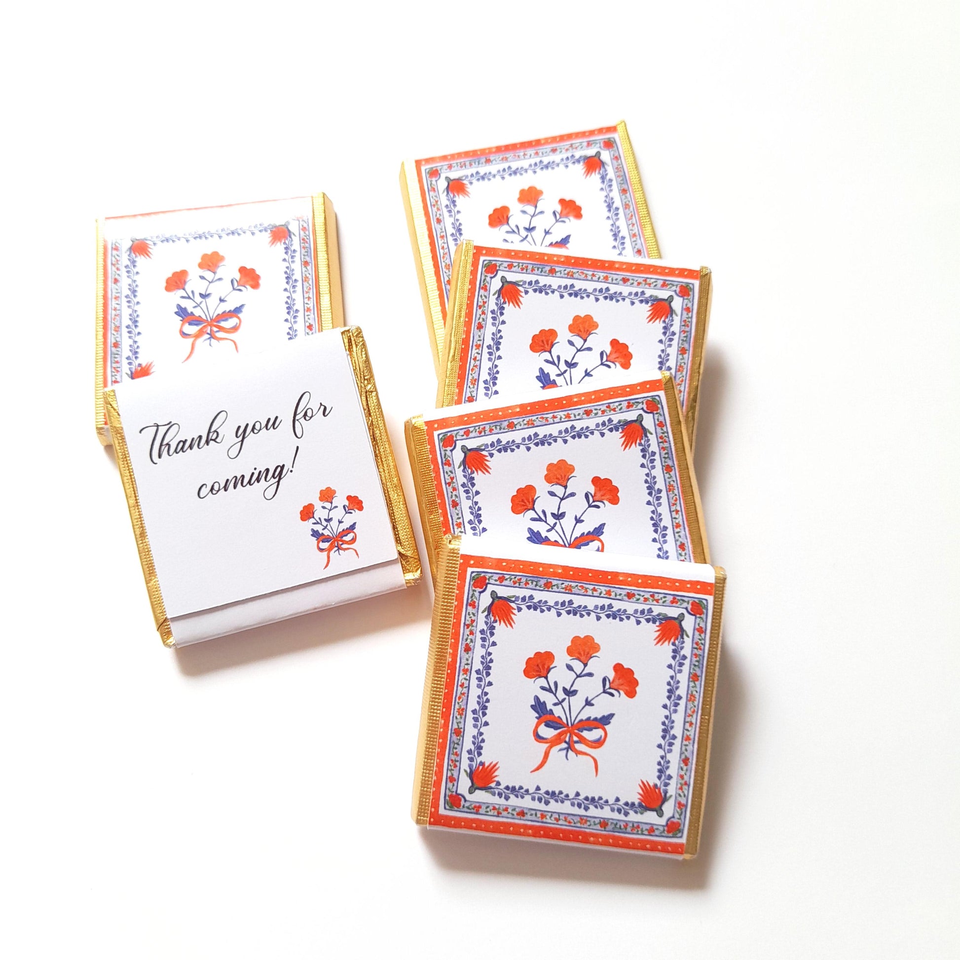 May include: Small chocolate bars with floral designs in red, blue, and white, with gold foil edges. One bar has a folded paper with the text "Thank you for coming!" and a matching floral design. The chocolates are arranged on a white surface.