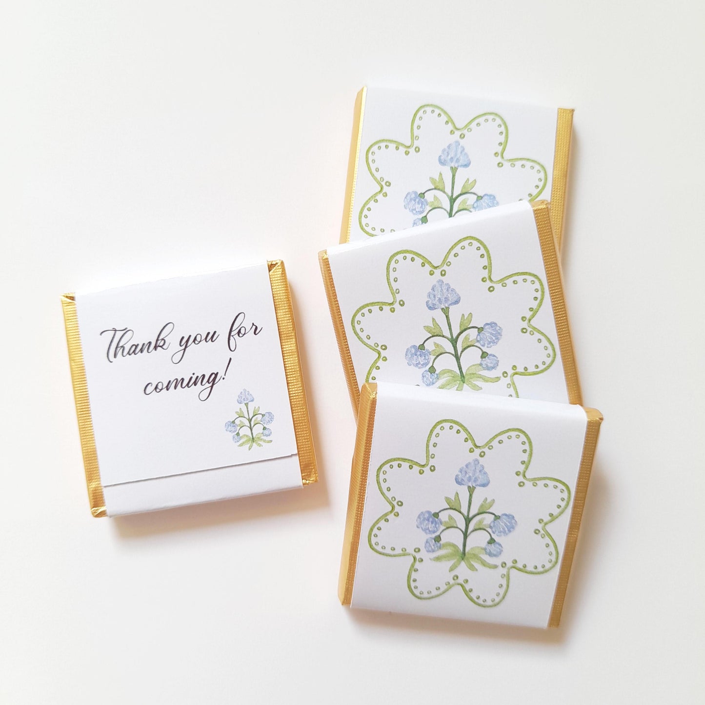 May include: Four small chocolate bars with gold foil edges. Three bars feature a floral design with blue flowers and green accents. One bar has the text "Thank you for coming!" in script.