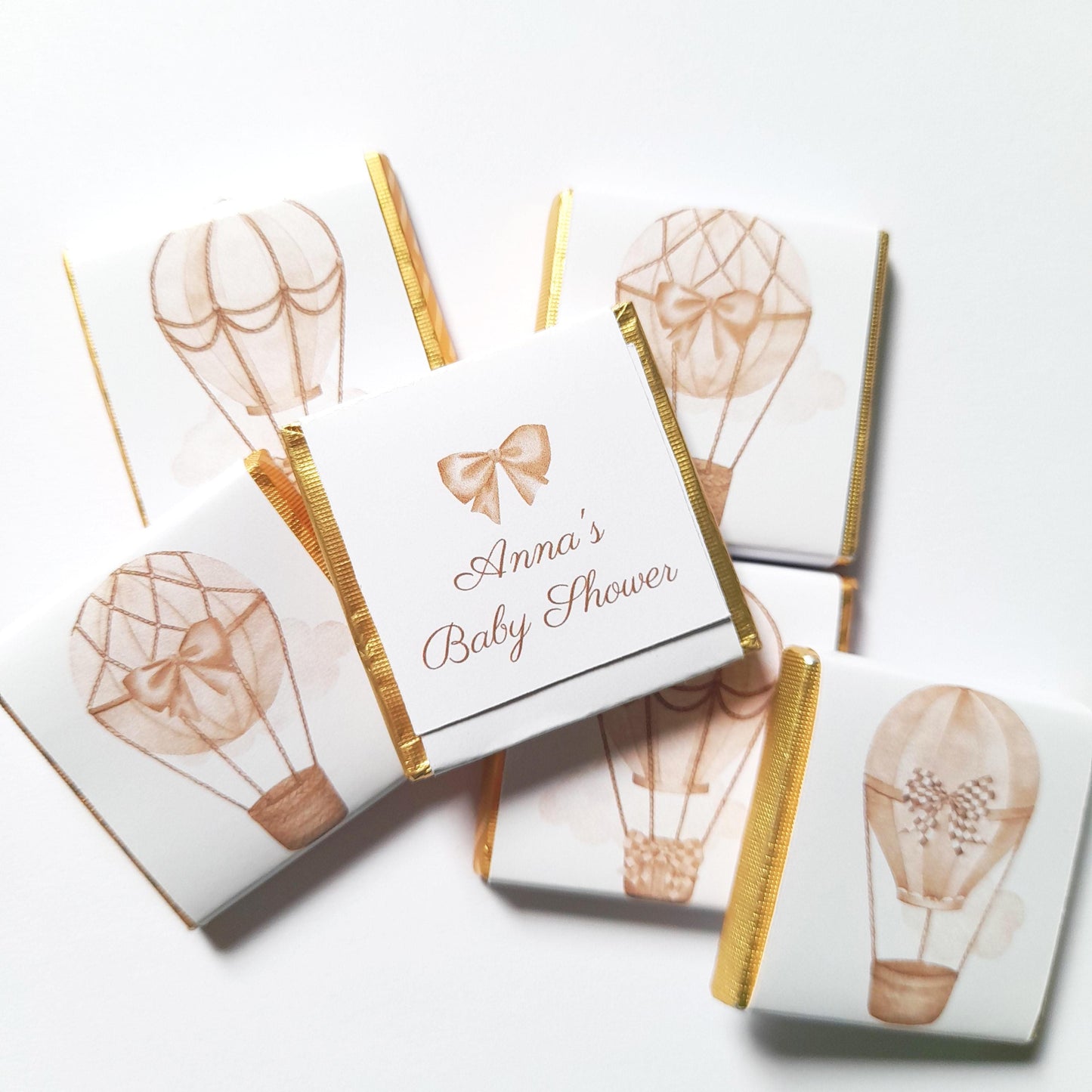 May include: Square white chocolates with gold foil edges, featuring watercolour hot air balloon illustrations in brown tones. One chocolate has the text "Anna's Baby Shower" with a bow graphic.