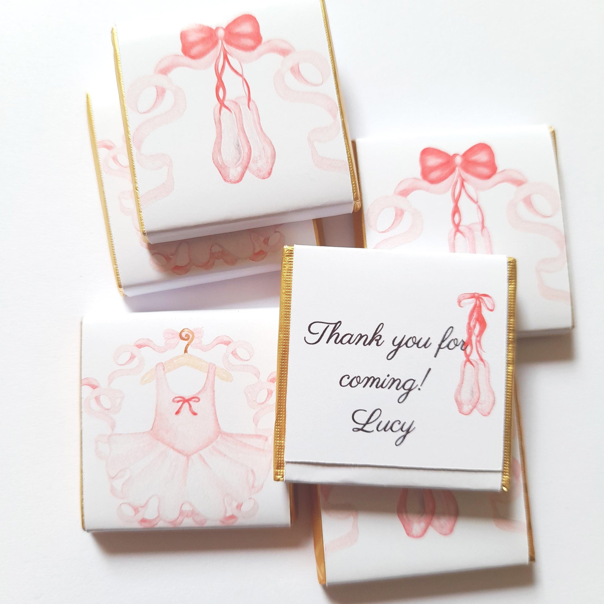 May include: Chocolate bar favours with pink ballet-themed illustrations. Some feature ballet shoes and ribbons, others a tutu on a hanger. One has the text "Thank you for coming! Lucy". Gold foil edges add elegance.