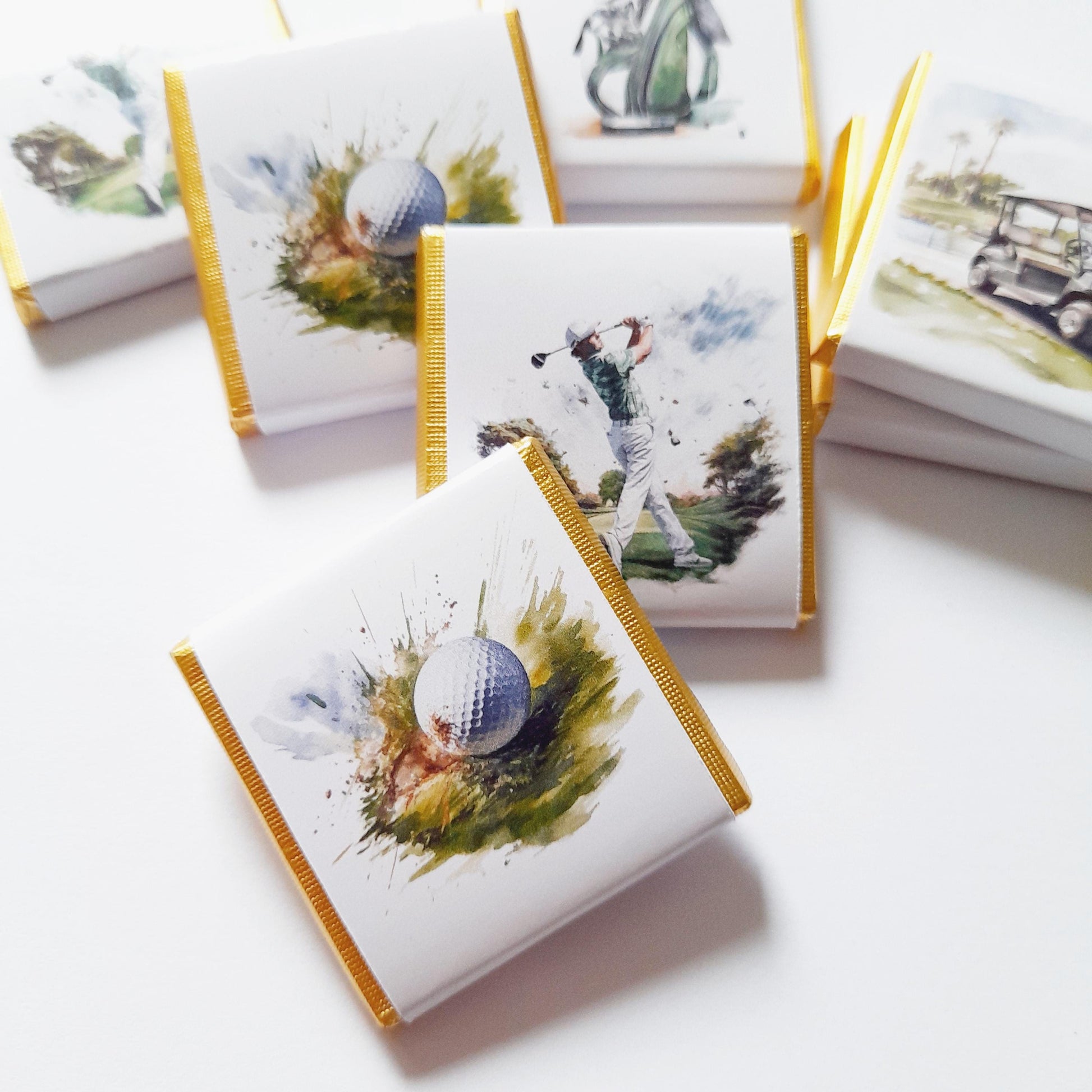 May include: A set of small, square, white chocolate bars with gold foil wrappers. Each bar features a different watercolour illustration of a golf-themed image, including a golfer swinging a club, a golf ball, and a golf cart.