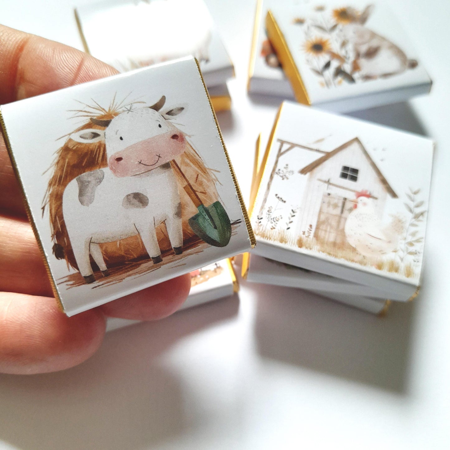 May include: A stack of small white boxes with gold trim, each featuring a different farm animal illustration. The top box shows a white cow with brown spots holding a green shovel, standing in a field of brown hay.
