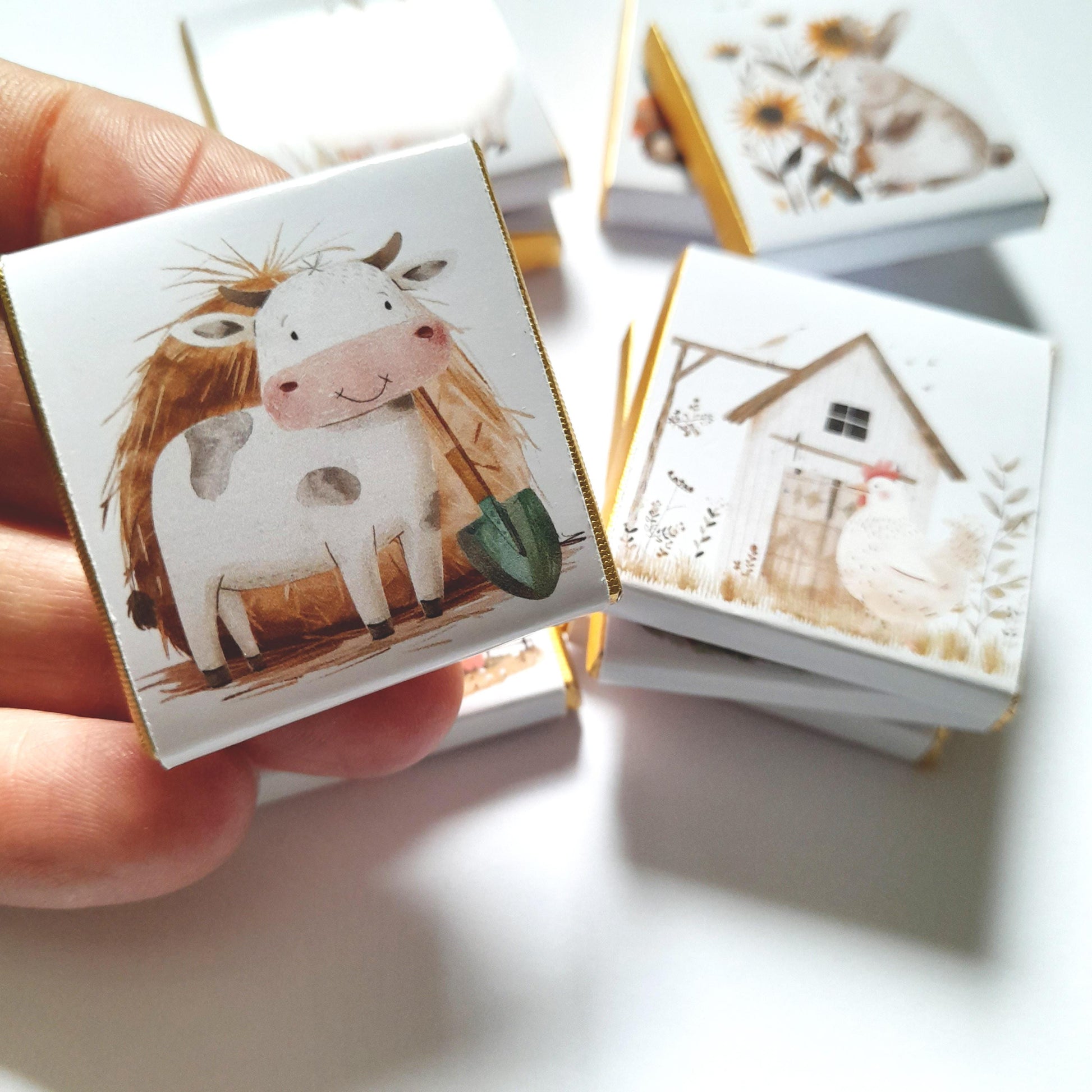 May include: A stack of small white boxes with gold trim, each featuring a different farm animal illustration. The top box shows a white cow with brown spots holding a green shovel, standing in a field of brown hay.