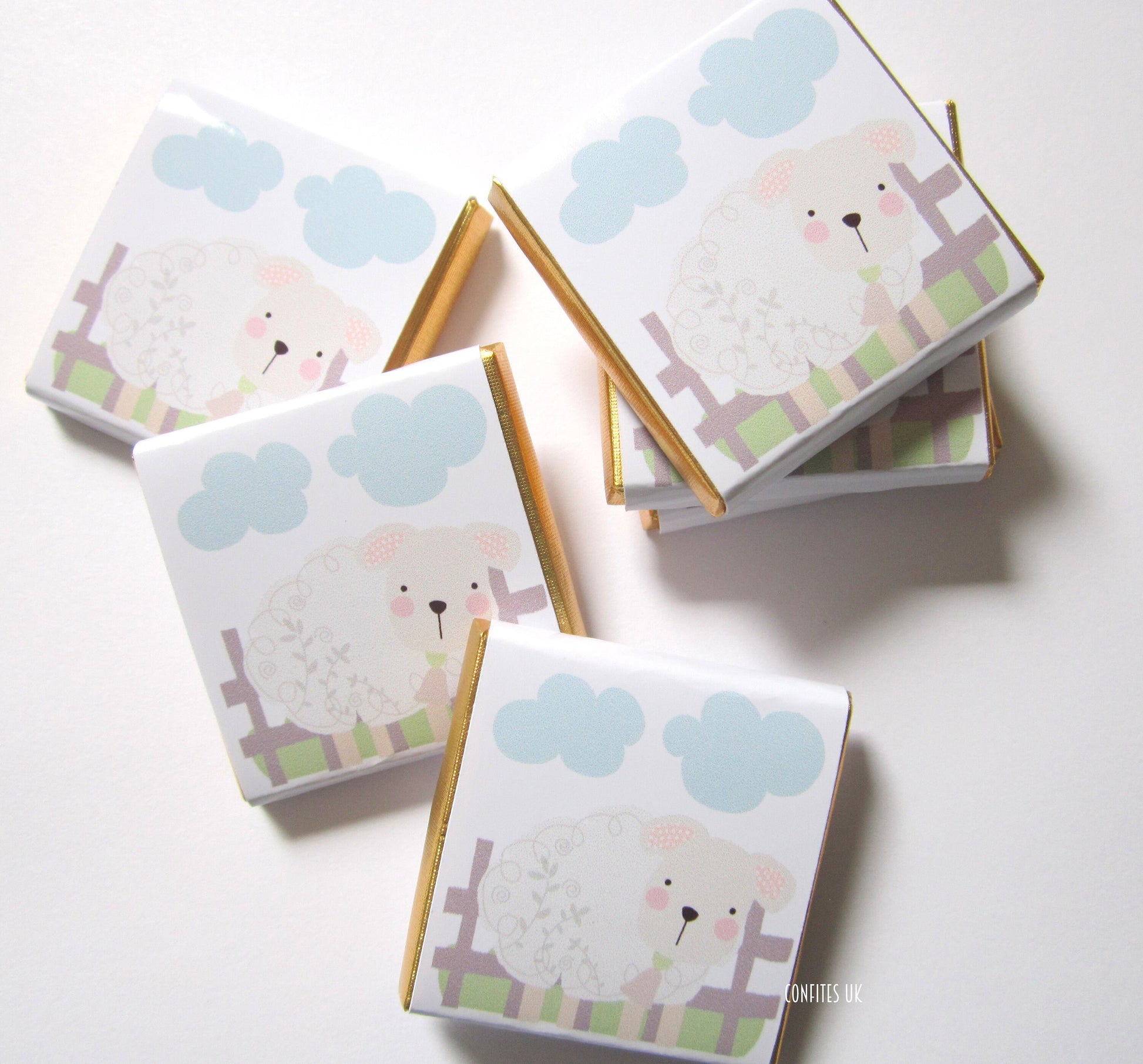 May include: A set of six white chocolate bars wrapped in gold foil. Each bar features a cute illustration of a sheep with blue clouds in the background. The text "Confites UK" is printed on one of the bars.