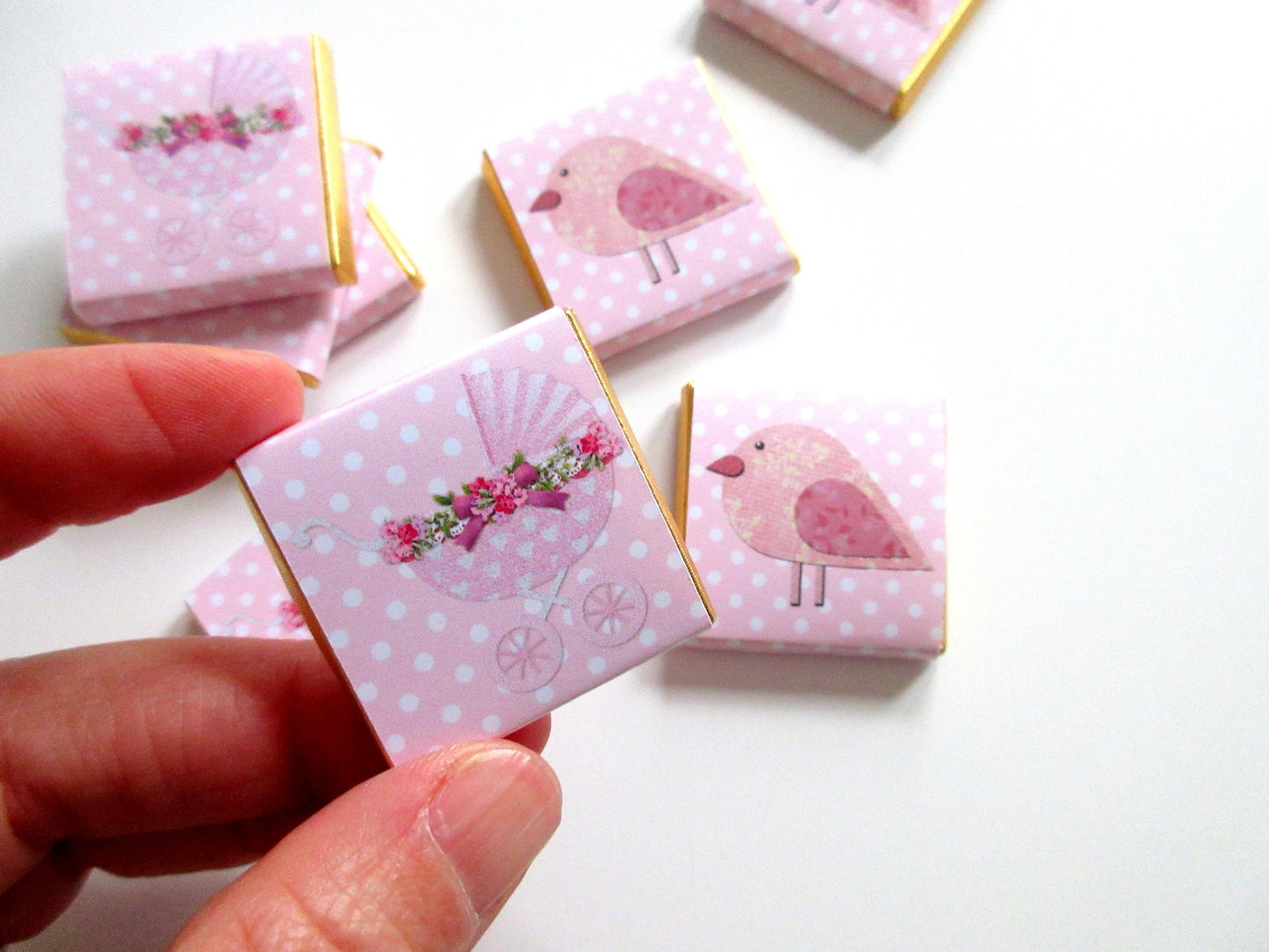 May include: Pink and white favour boxes with a gold trim. The boxes feature a pink bird and a pink baby carriage with white polka dots.