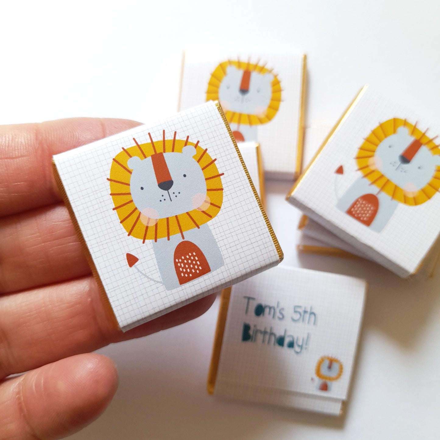May include: A stack of small, white boxes with a gold trim. Each box features a cartoon lion with a yellow mane and a white background. The top box has the text "Tom's 5th Birthday!" printed on it.