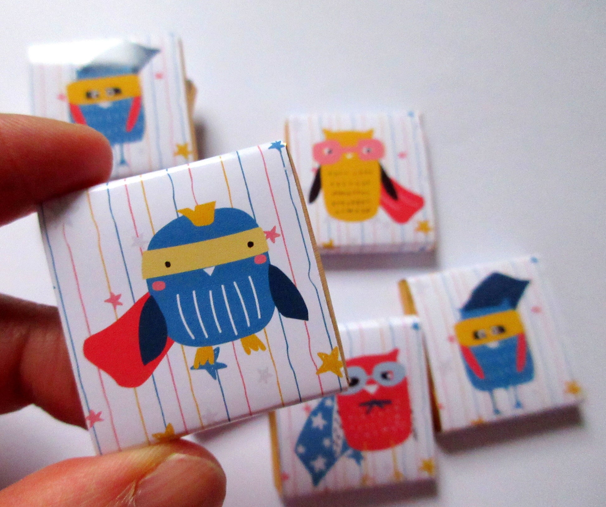 May include: A selection of square magnets with cartoon owl superhero designs. Each magnet showcases a different owl character with capes, masks, and star details. The background features vertical stripes and small stars.