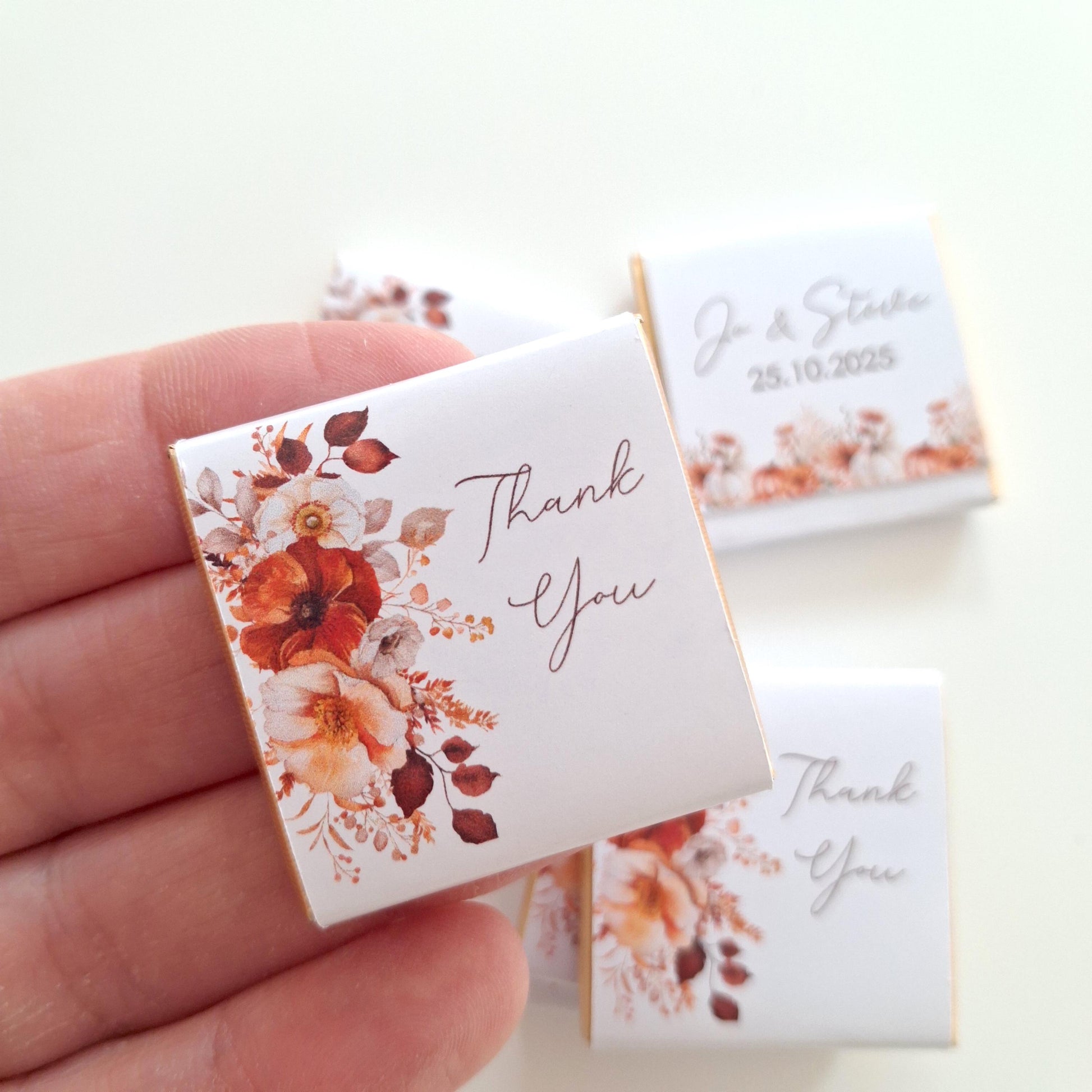 May include: Square white chocolate favours with floral designs in shades of orange, red, and cream. The words "Thank You" are printed on the front. Some favours have names and a date printed on them. The favours are held in a hand.