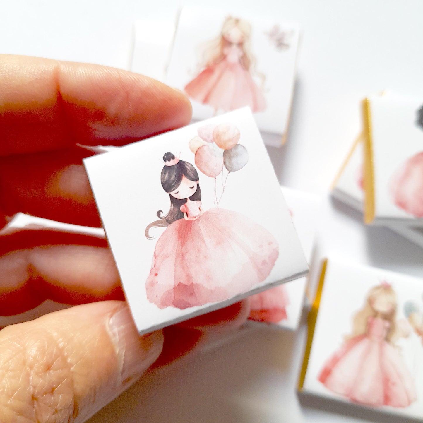 May include: A small white box with a pink and white illustration of a girl in a pink dress holding balloons. The girl has brown hair and a crown on her head. The balloons are pink, white, and grey.