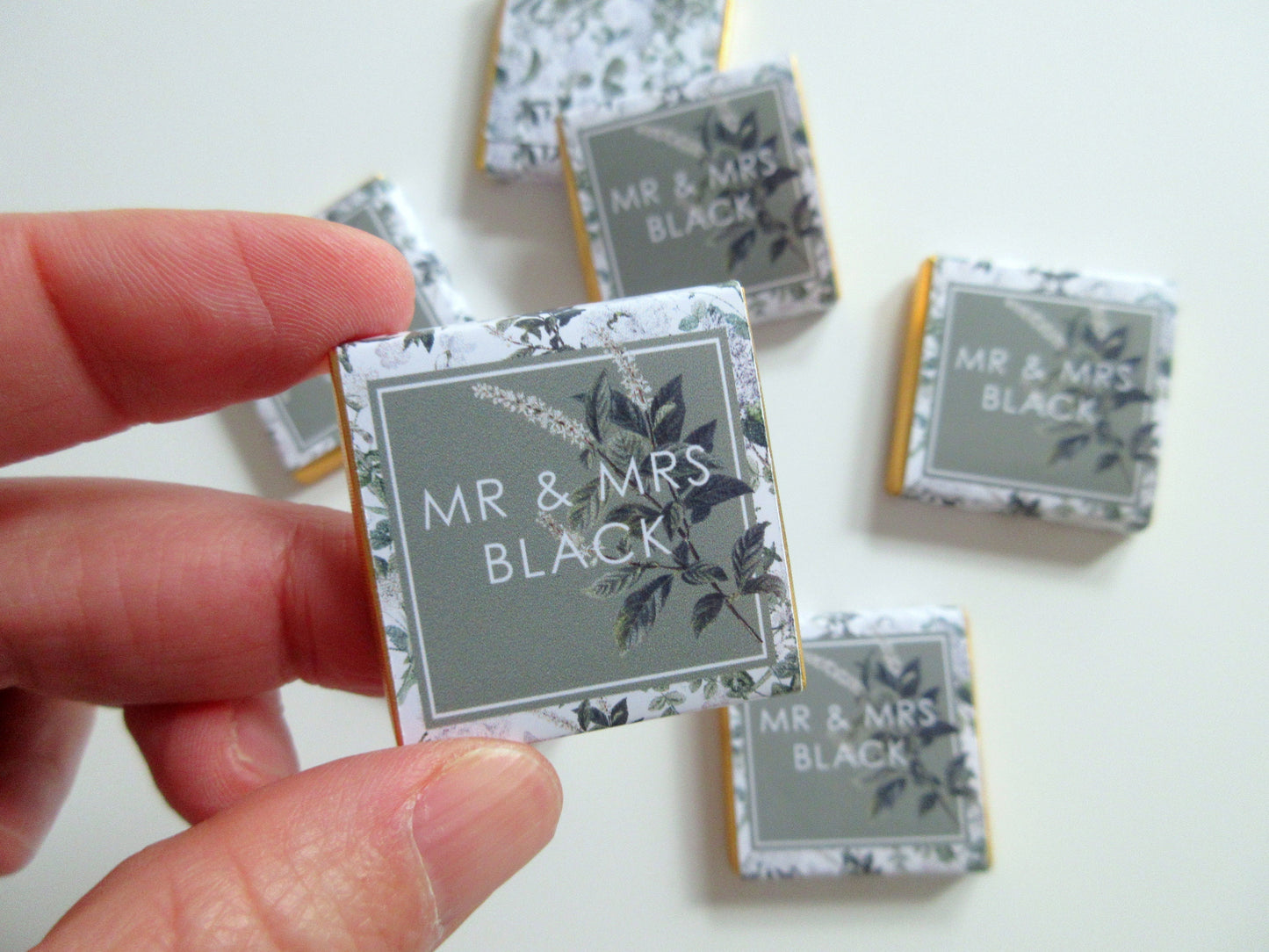May include: A set of small, square, chocolate favours with a gold border and a white background. The favours are decorated with a green and white floral design and the text "MR & MRS BLACK".
