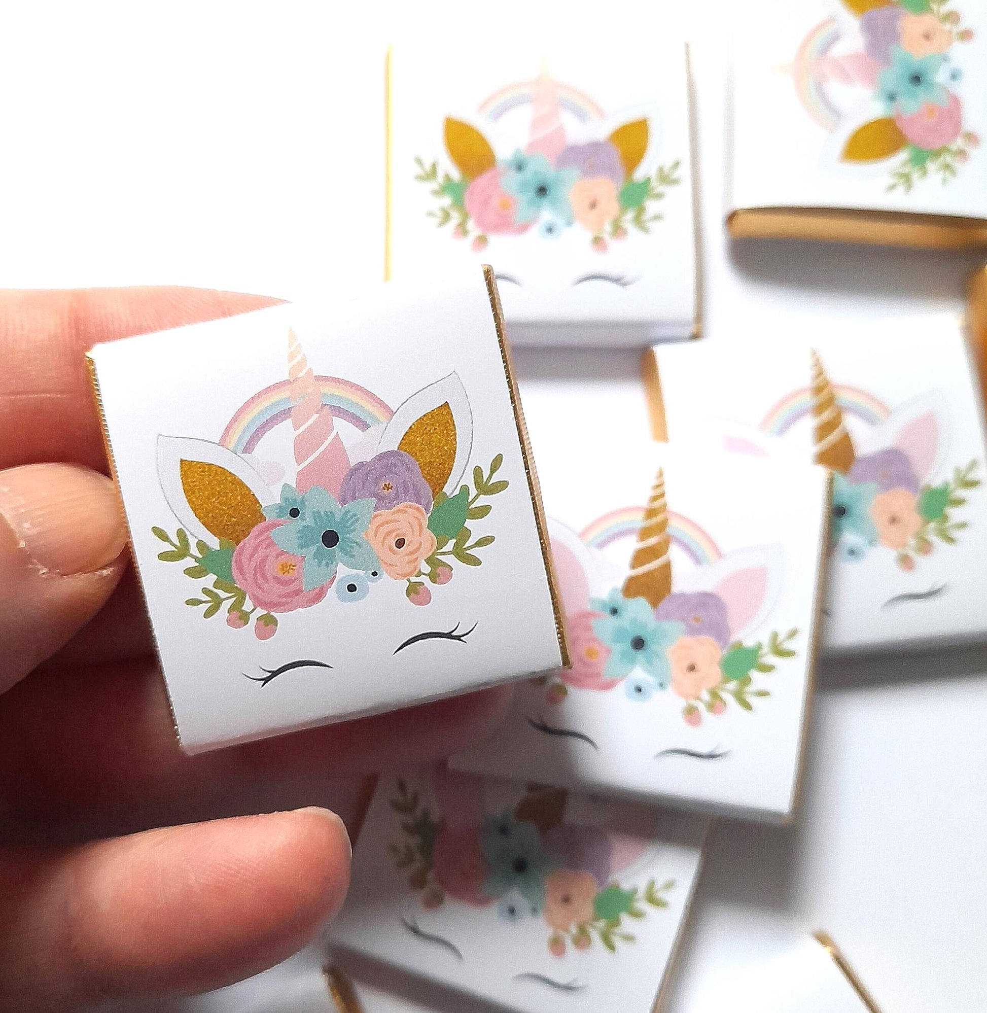 May include: A set of small white chocolate bars with gold foil wrappers. Each bar features a colourful illustration of a unicorn with a rainbow, flowers, and a gold horn.