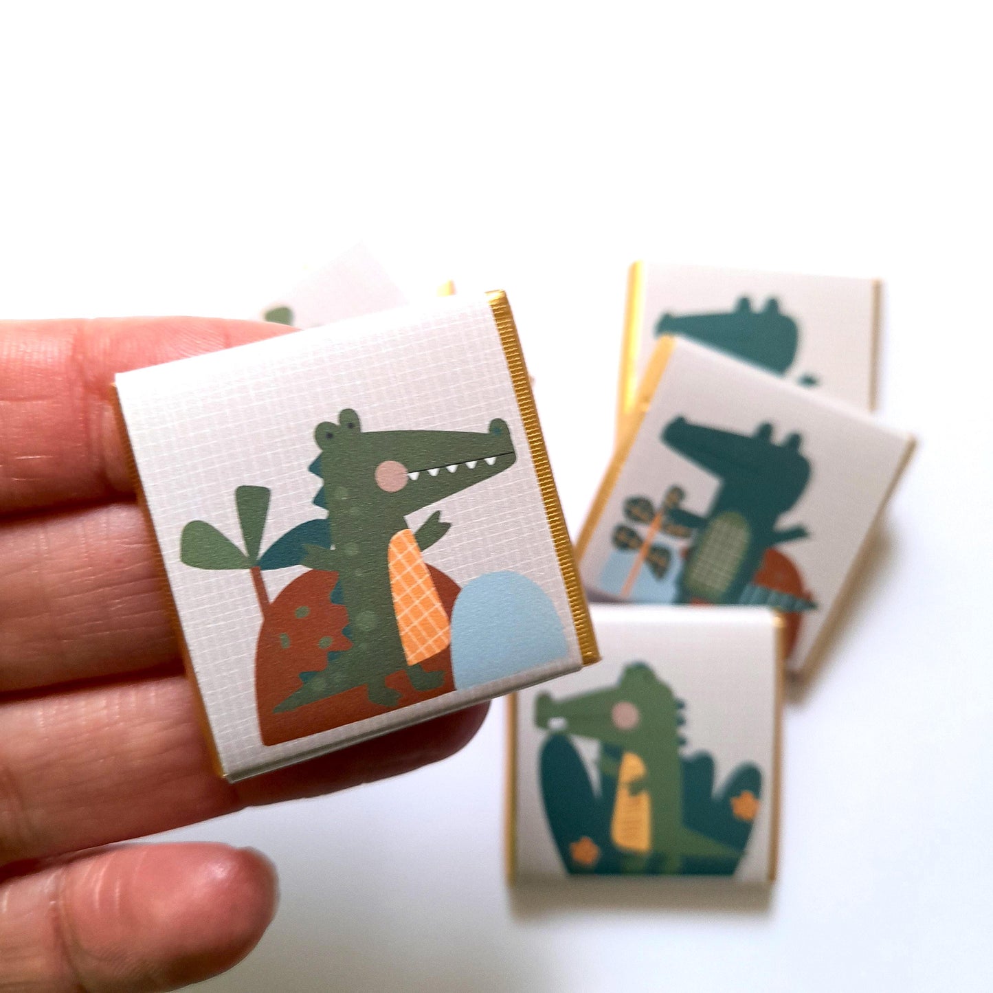 May include: A set of small, white chocolate bars with gold foil accents. Each bar features a different illustration of a green crocodile with a cartoonish style. The crocodile illustrations are set against a background of brown, orange, and blue.
