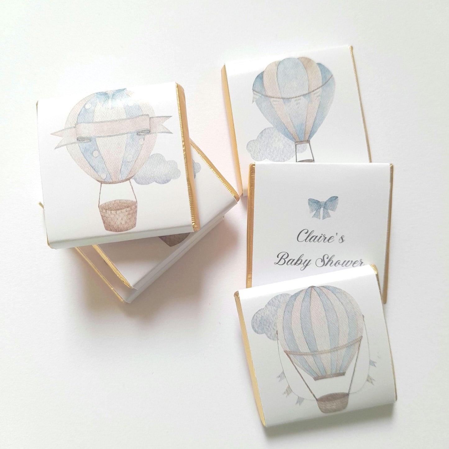 May include: A stack of white chocolate bar favours with gold foil accents. Each bar features a watercolour illustration of a blue and white hot air balloon with a basket. One bar has the text "Claire's Baby Shower" printed on it.