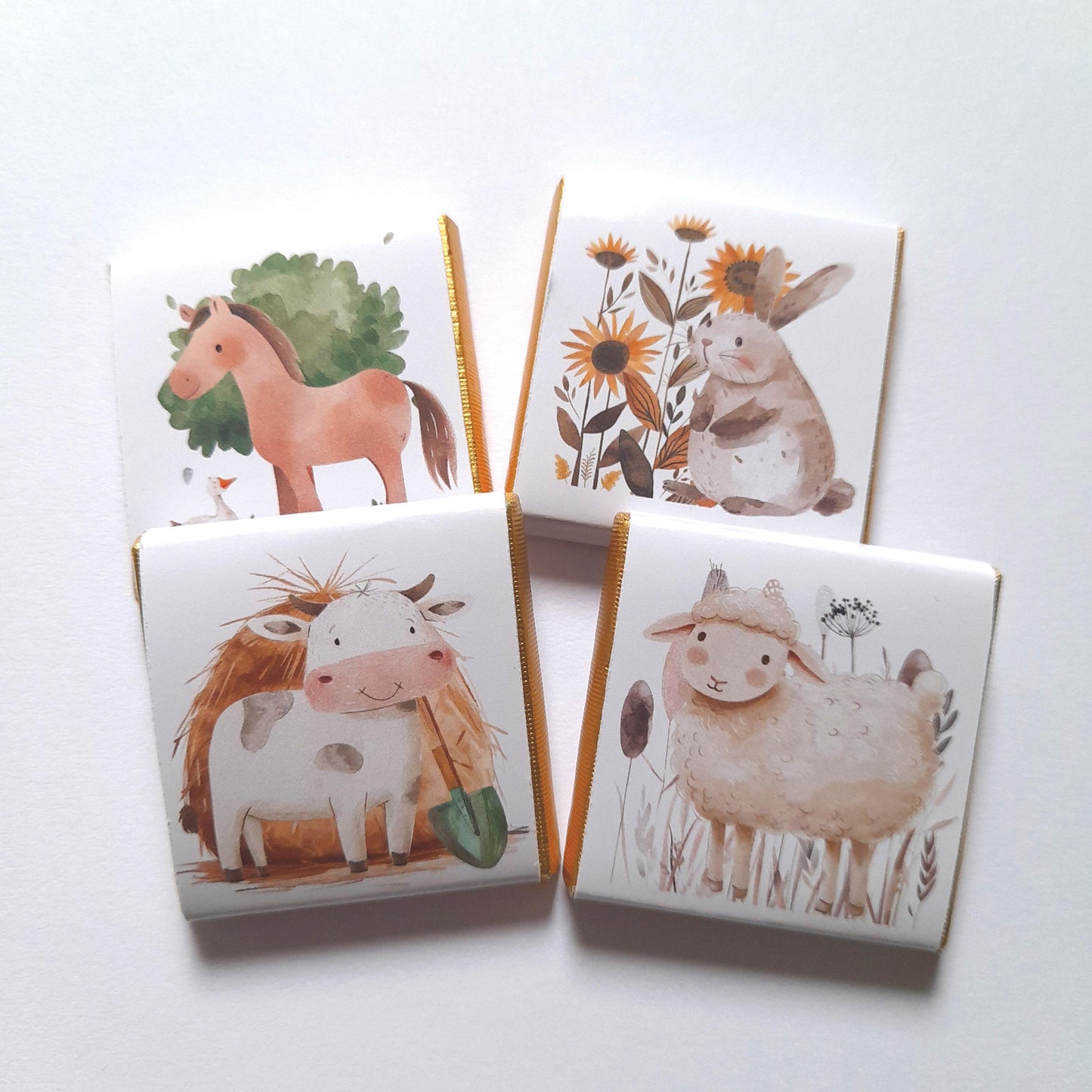 May include: Four small white chocolate bars with gold foil trim. Each bar features a different farm animal illustration: a horse, a cow, a sheep, and a rabbit. The rabbit is surrounded by flowers.