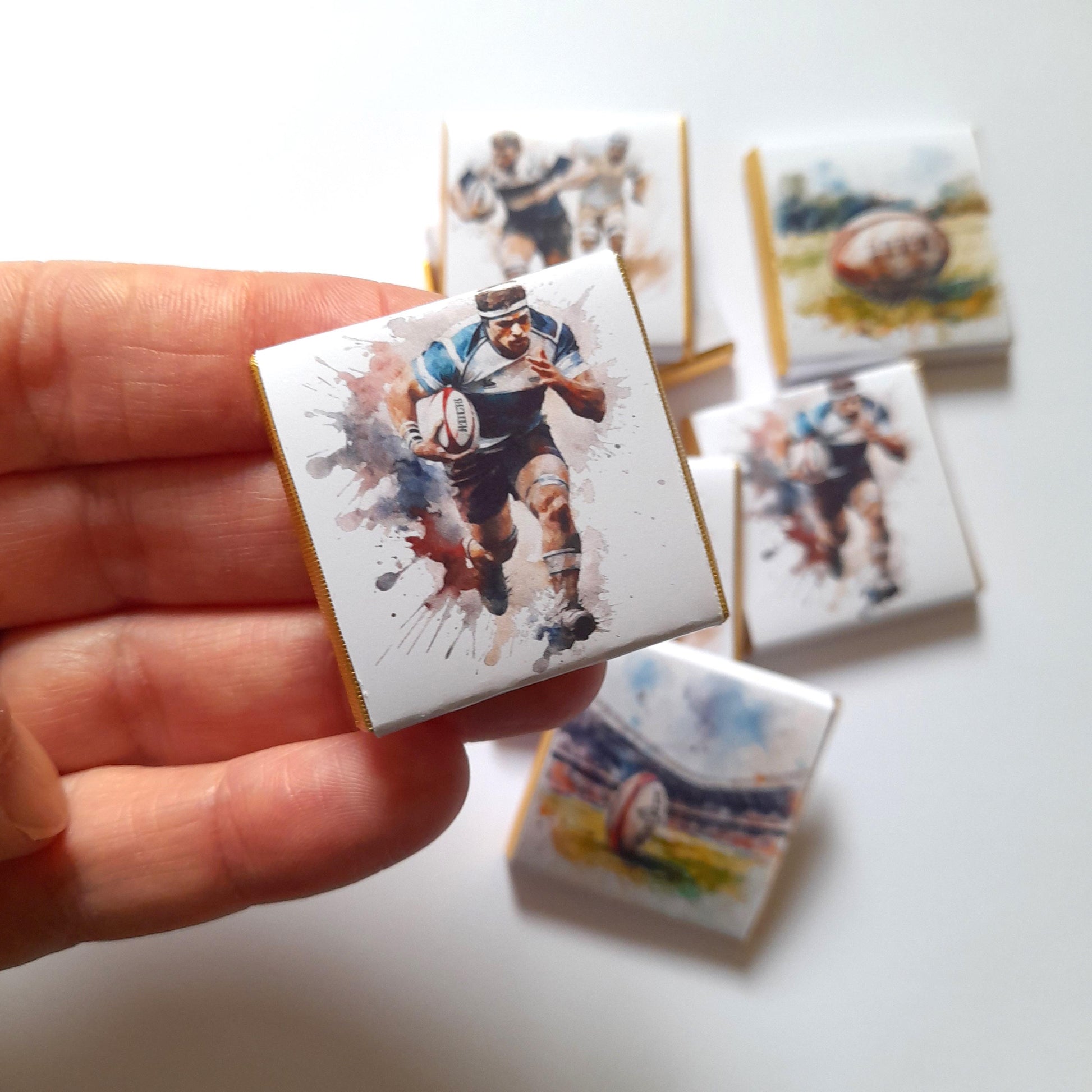May include: A set of six small chocolate bars with rugby-themed designs. The bars are wrapped in gold foil and feature watercolour illustrations of rugby players and a rugby ball.