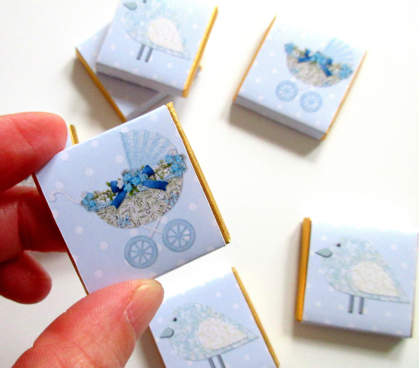 May include: Blue and white baby shower favours with gold trim. The favours feature a baby carriage with blue and white flowers and a blue bird.