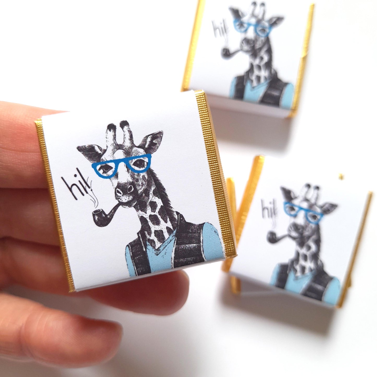 May include: Three small white boxes with gold trim, each featuring a cartoon giraffe wearing blue glasses and a pipe. The boxes have the word "hil" printed on them.