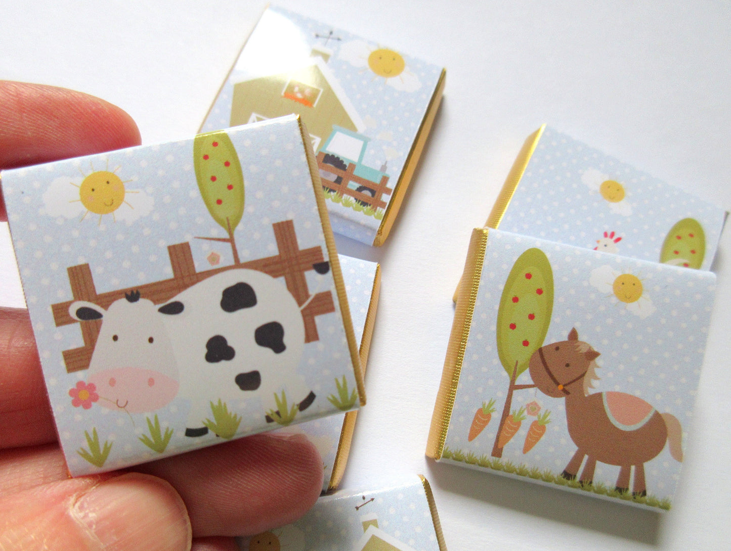 May include: Square chocolates with farm animal illustrations. Each wrapper shows a different farm scene: a cow, a horse, a barn, and a tractor. The wrappers have a light blue background with white polka dots and a gold border.