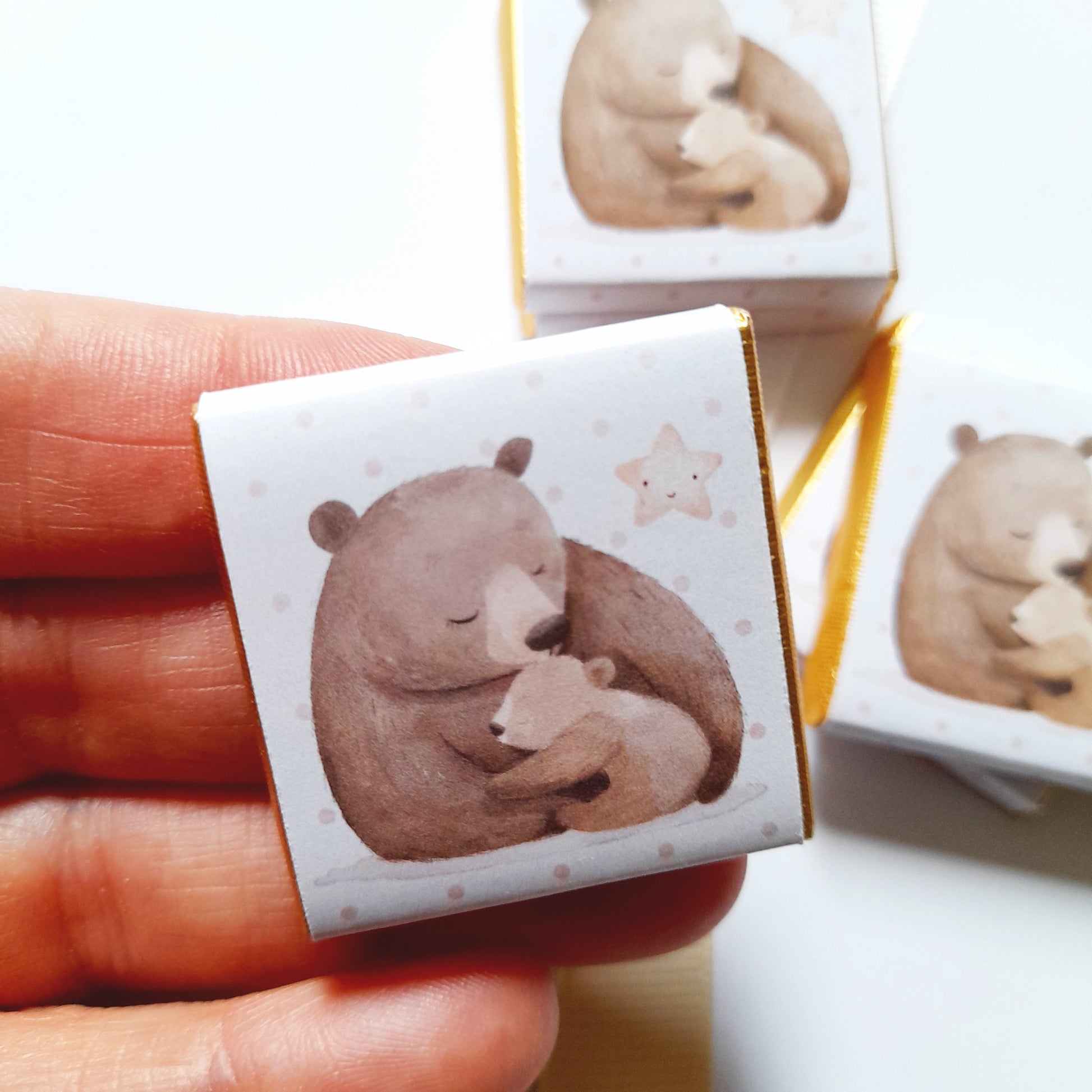 May include: A set of three small, white gift boxes with gold trim. Each box features a watercolour illustration of a brown bear hugging a smaller bear cub. The background is a light pink with white polka dots and a gold star.