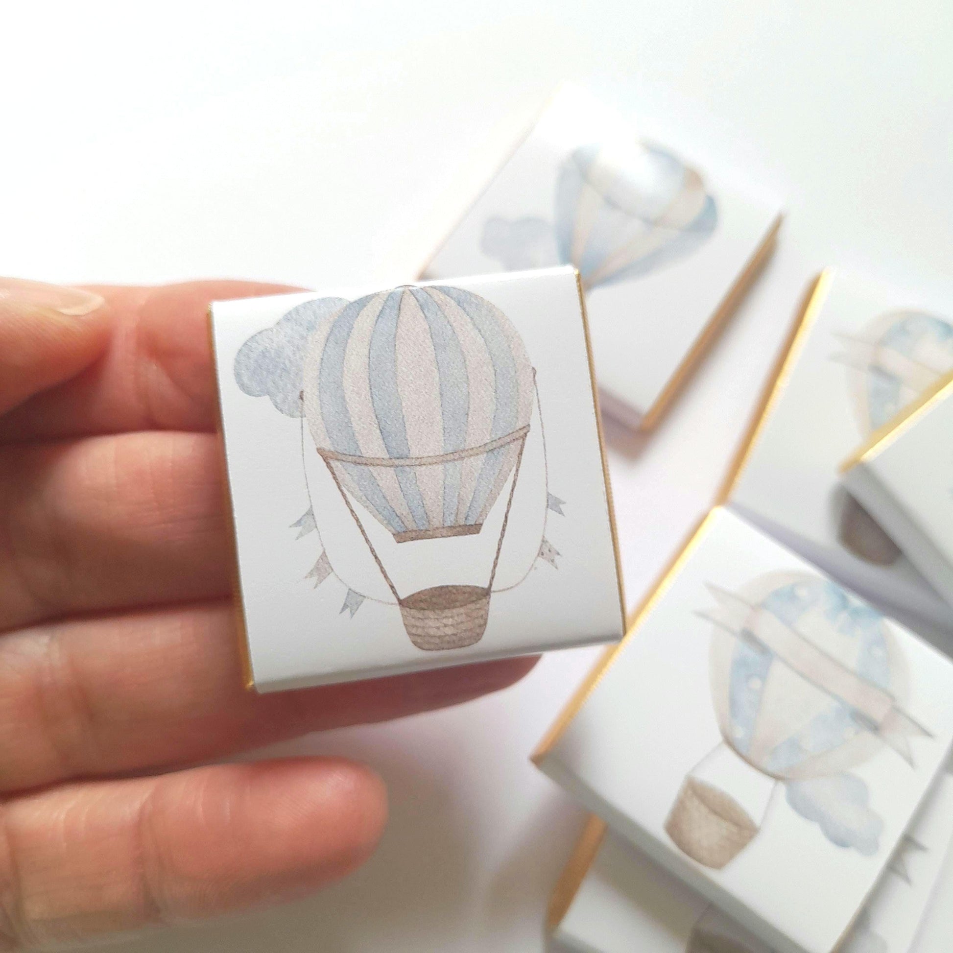 May include: A set of small, white, square boxes with gold trim. Each box features a watercolour illustration of a blue and white hot air balloon with a basket. The boxes are ideal for favours or gifts.
