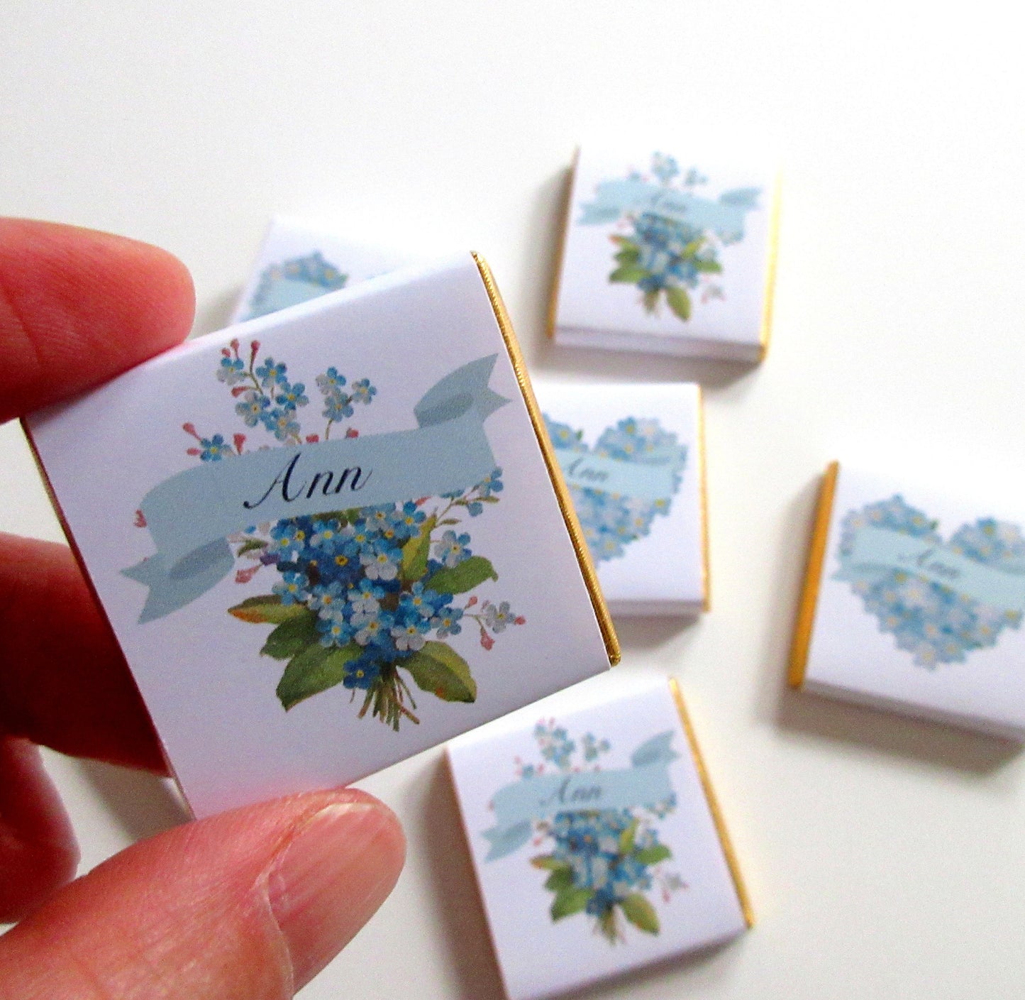May include: Five small, square, white chocolate boxes with gold foil trim. Each box has a blue and white floral design with a blue ribbon and the name "Ann" printed on the ribbon.