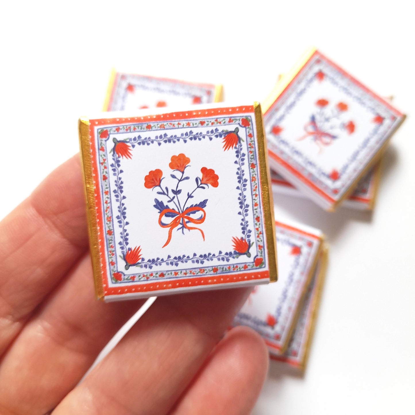 May include: Small, square chocolates with floral designs. Each chocolate has a white background with a red and blue floral pattern and a gold foil border. The chocolates are held in a hand.