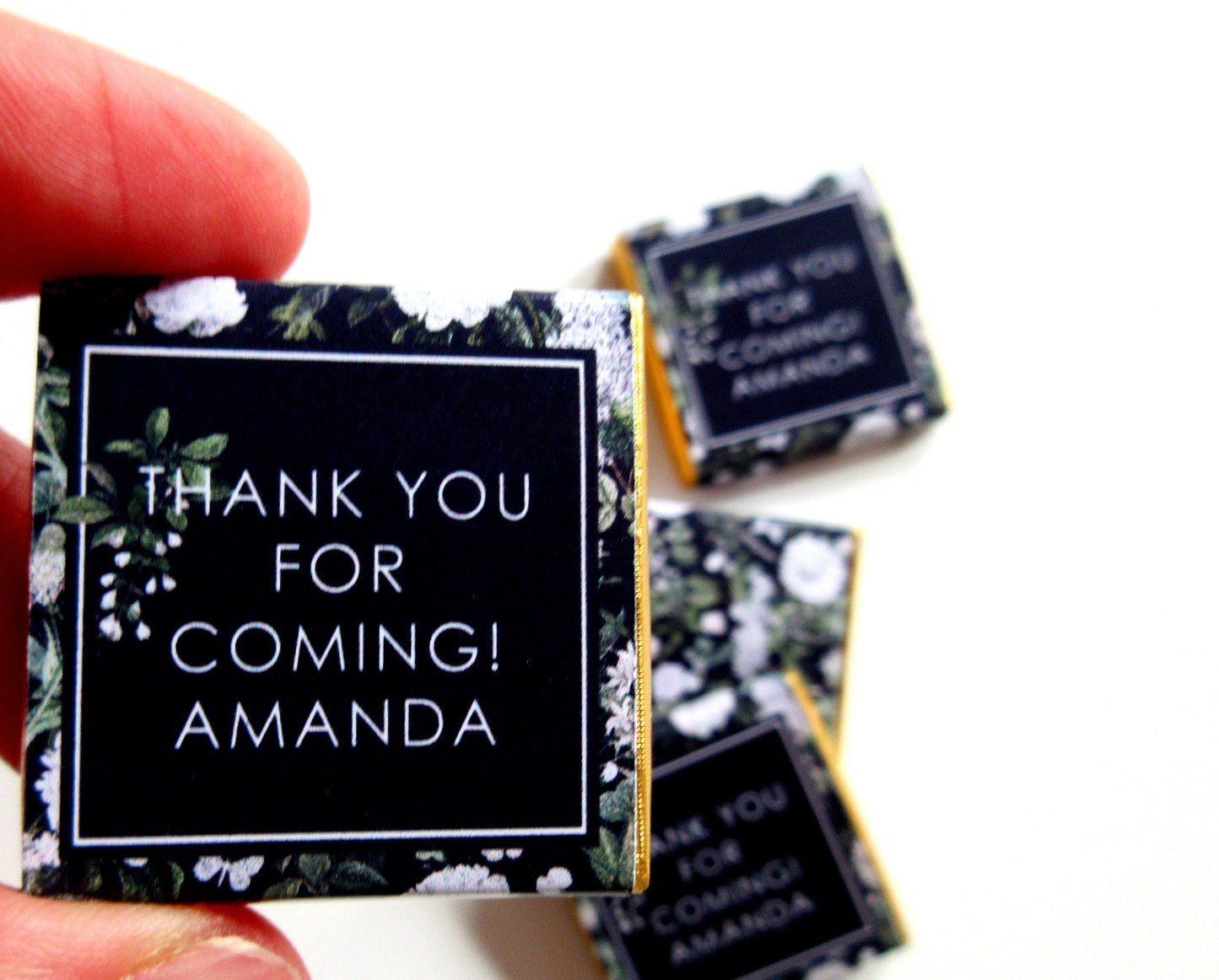 May include: Miniature chocolate favors with a black background and a white floral pattern. The text "THANK YOU FOR COMING! AMANDA" is printed in white. Several favors are visible, some held by a person.