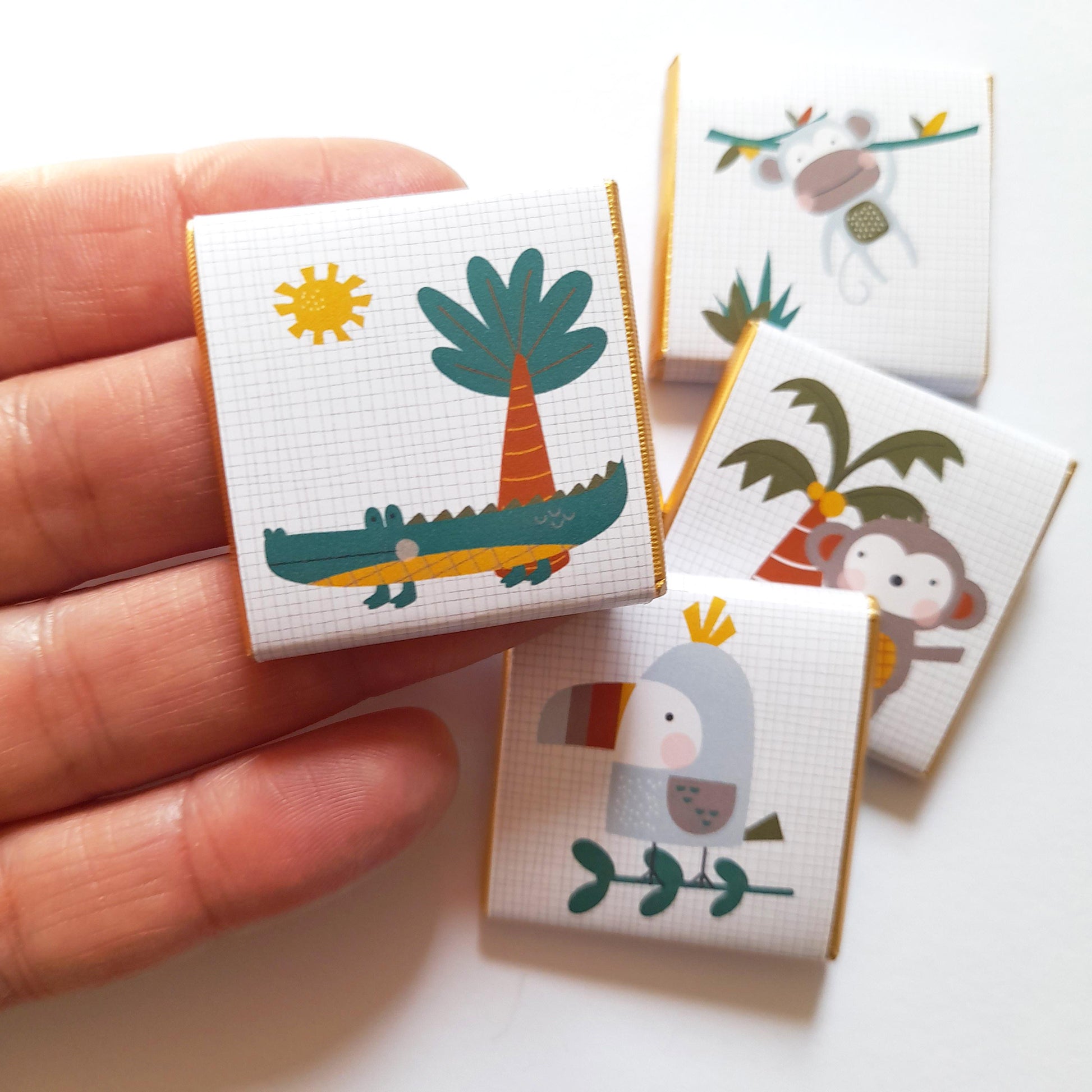 May include: Small, square chocolate bars featuring jungle animal designs. The illustrations include a crocodile, monkey, toucan, and palm trees on a white grid background. Each bar has gold foil edging.