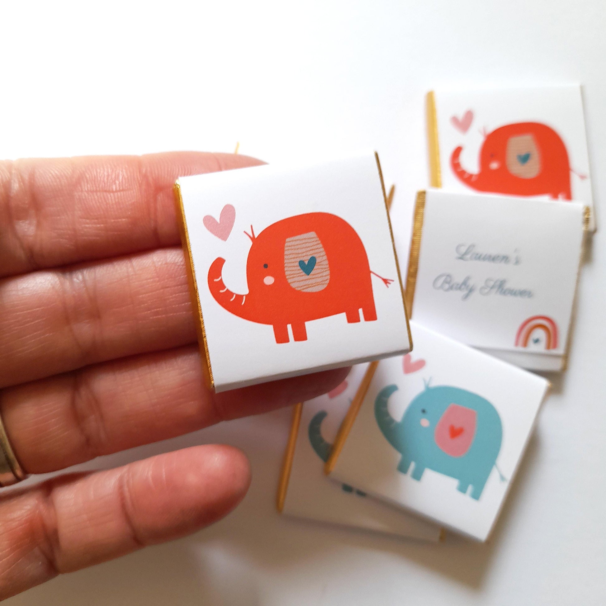 May include: A set of small, white favour boxes with gold trim. Each box features a cartoon elephant with a pink heart on its chest. The boxes are decorated with a pink heart and the text "Lauren's Baby Shower".