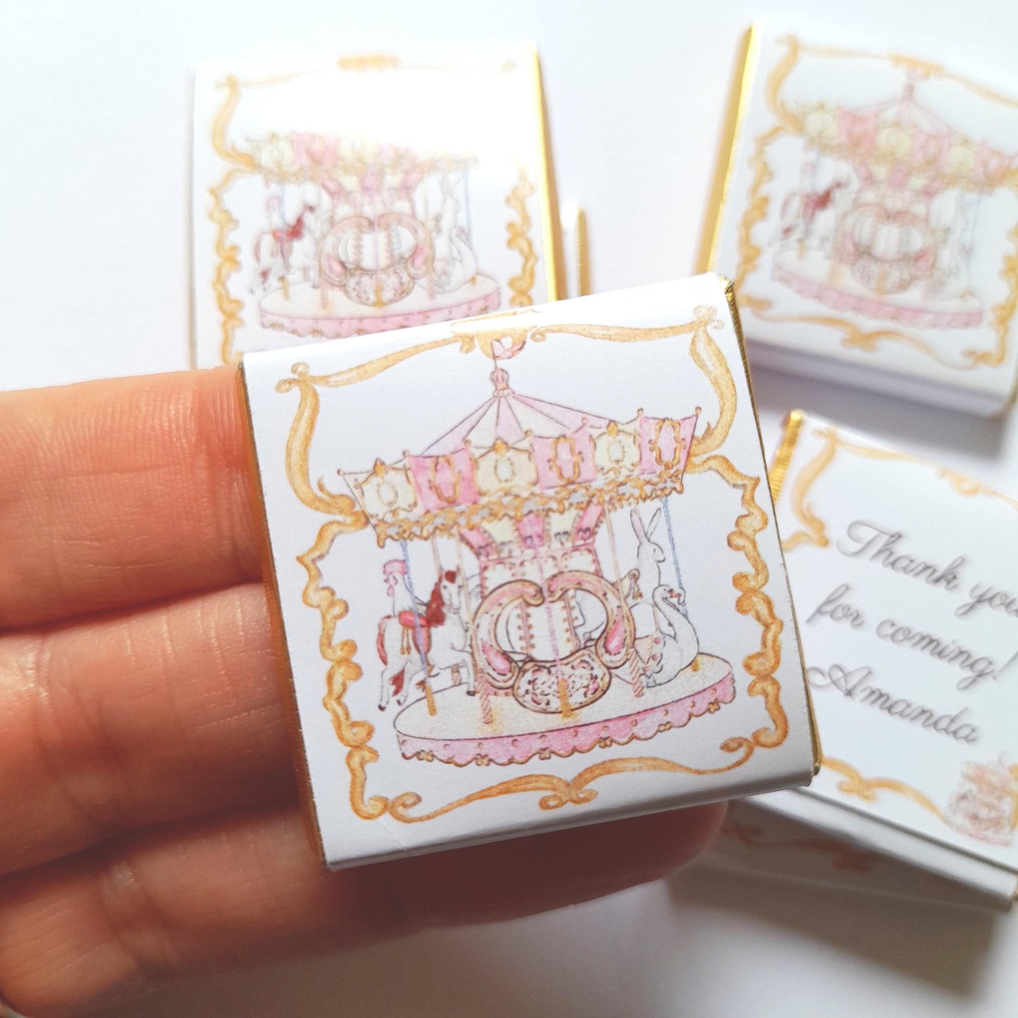 May include: Small, square favour boxes with a carousel design. The boxes feature a pink and gold carousel illustration on a white background. One box has the text "Thank you for coming! Amanda".
