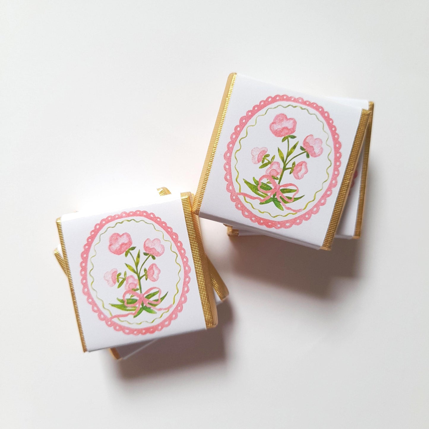 May include: Square white chocolate bars with gold foil edges. Each bar features a pink floral design within a scalloped pink and green oval. The floral design includes pink flowers, green leaves, and a pink bow.