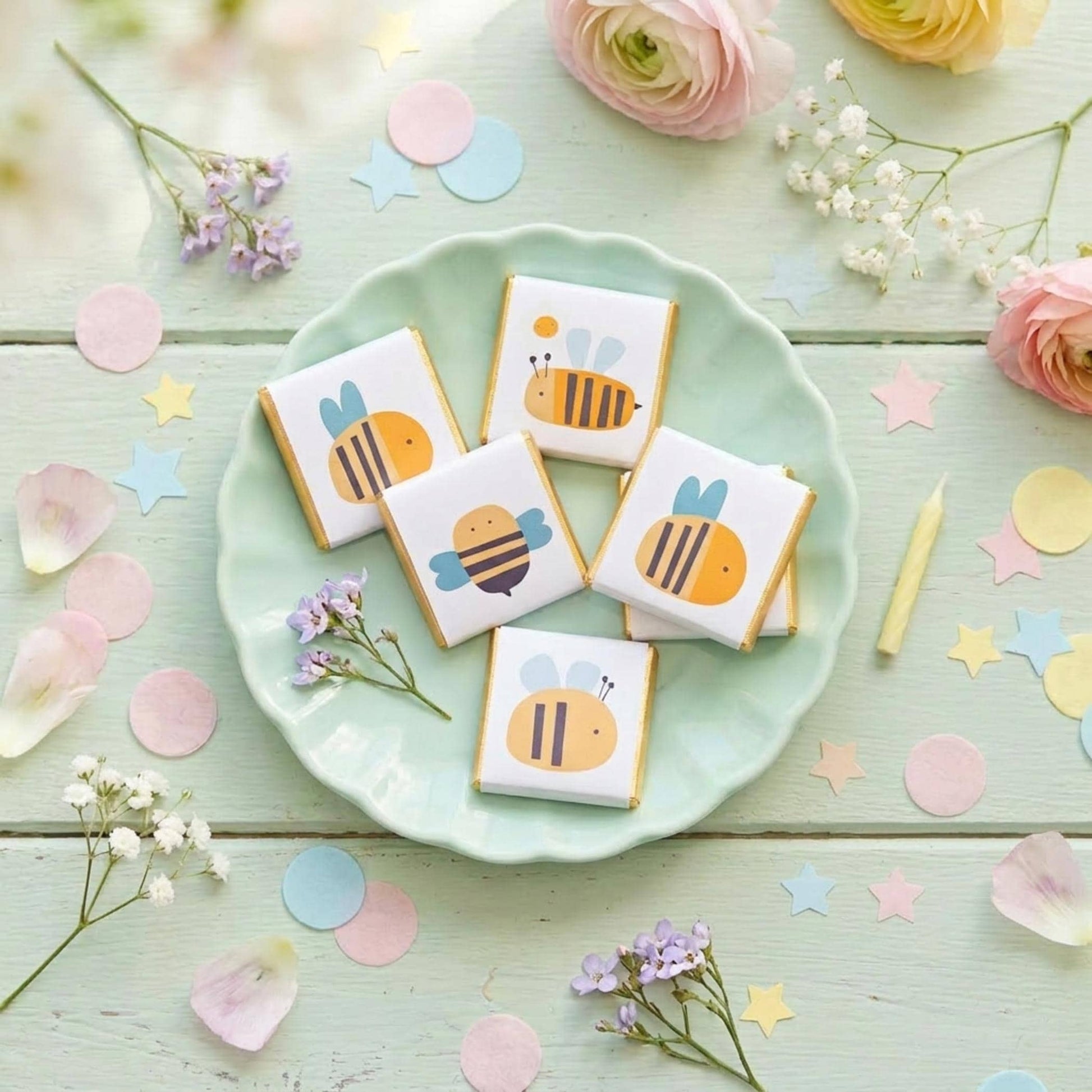 May include: Five square chocolates with bee designs sit on a light green plate. The bees are yellow, orange, and blue, with gold foil edges. The image is decorated with flowers, confetti, and a yellow candle.