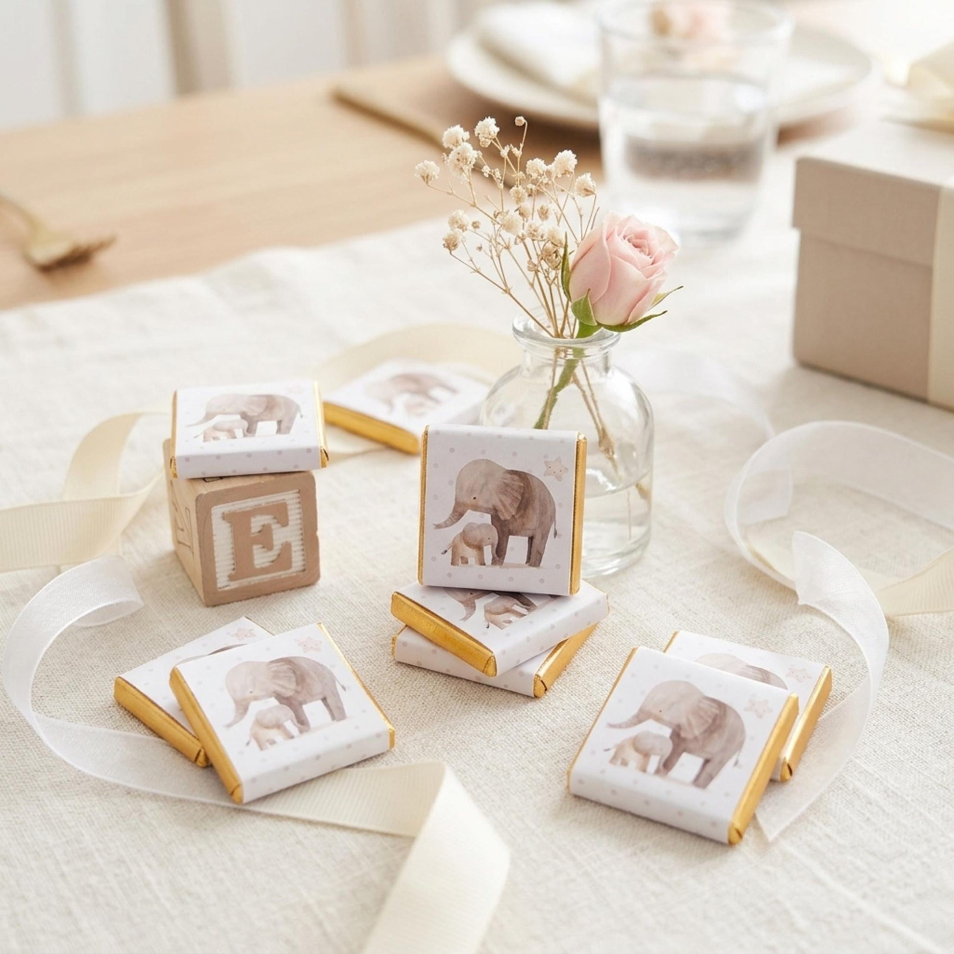May include: Small square chocolates with gold foil edges, each displaying an elephant and calf illustration. A wooden block with the letter 'E' is visible. The arrangement includes a small vase with a pink rose and white ribbon.
