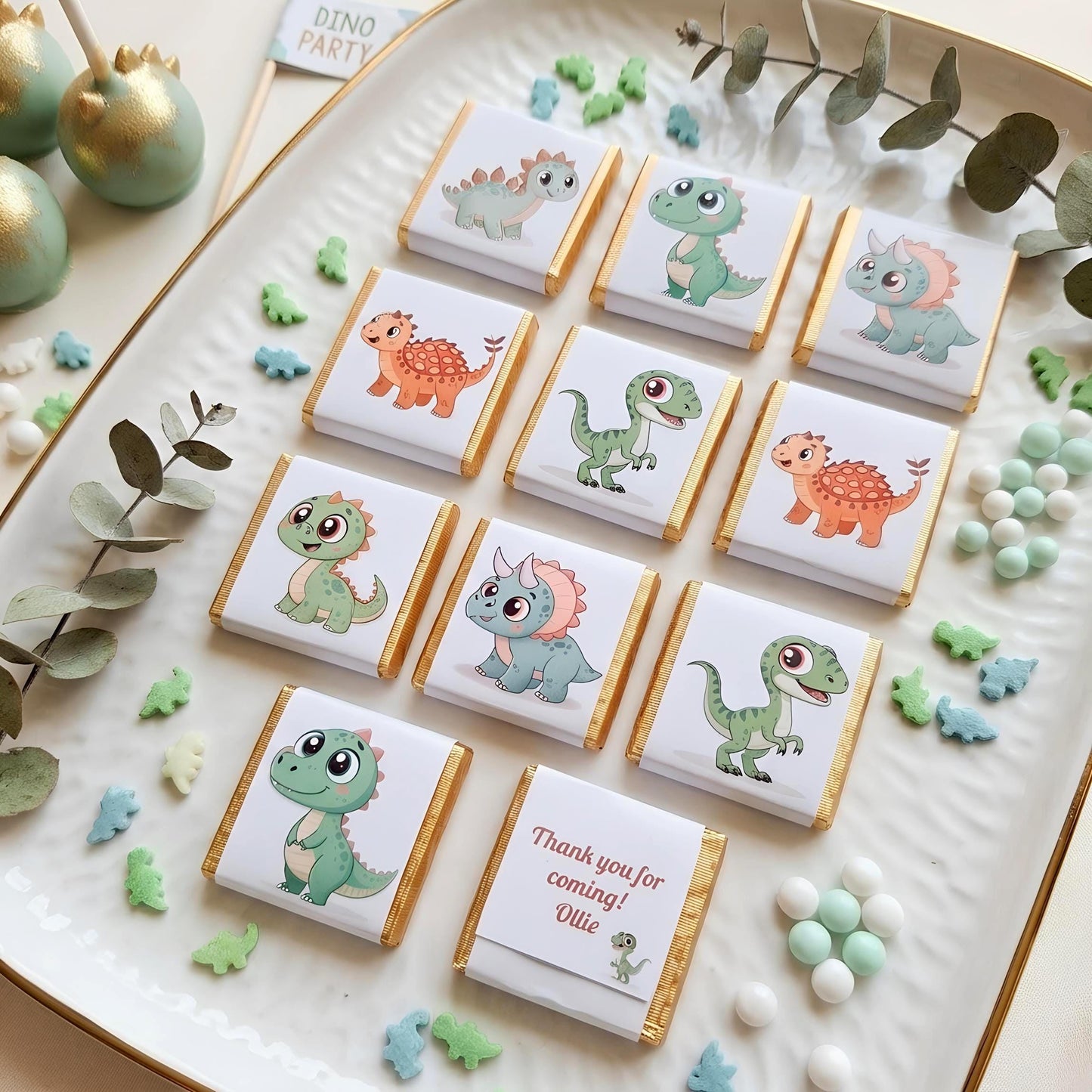 May include: A white serving platter displays chocolate bars with dinosaur designs. The bars have gold foil edges and feature cartoon dinosaurs. Some bars have the text "Thank you for coming! Ollie". Green and blue dinosaur-shaped sprinkles are scattered around.