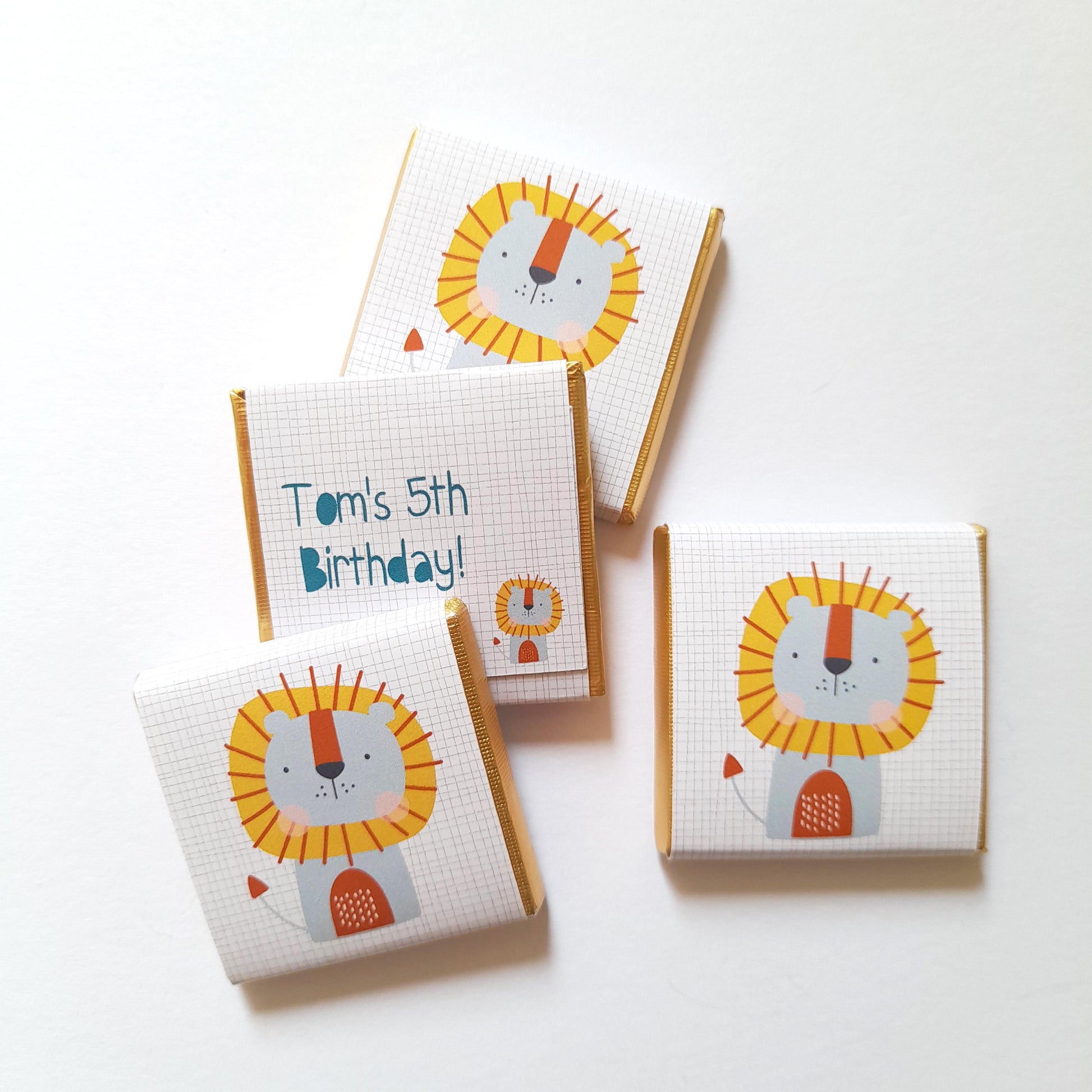 May include: Four small chocolate bars wrapped in white paper with a gold border. Each bar features a cartoon lion with a yellow mane and a blue face. One bar has the text "Tom's 5th Birthday!" printed on it.
