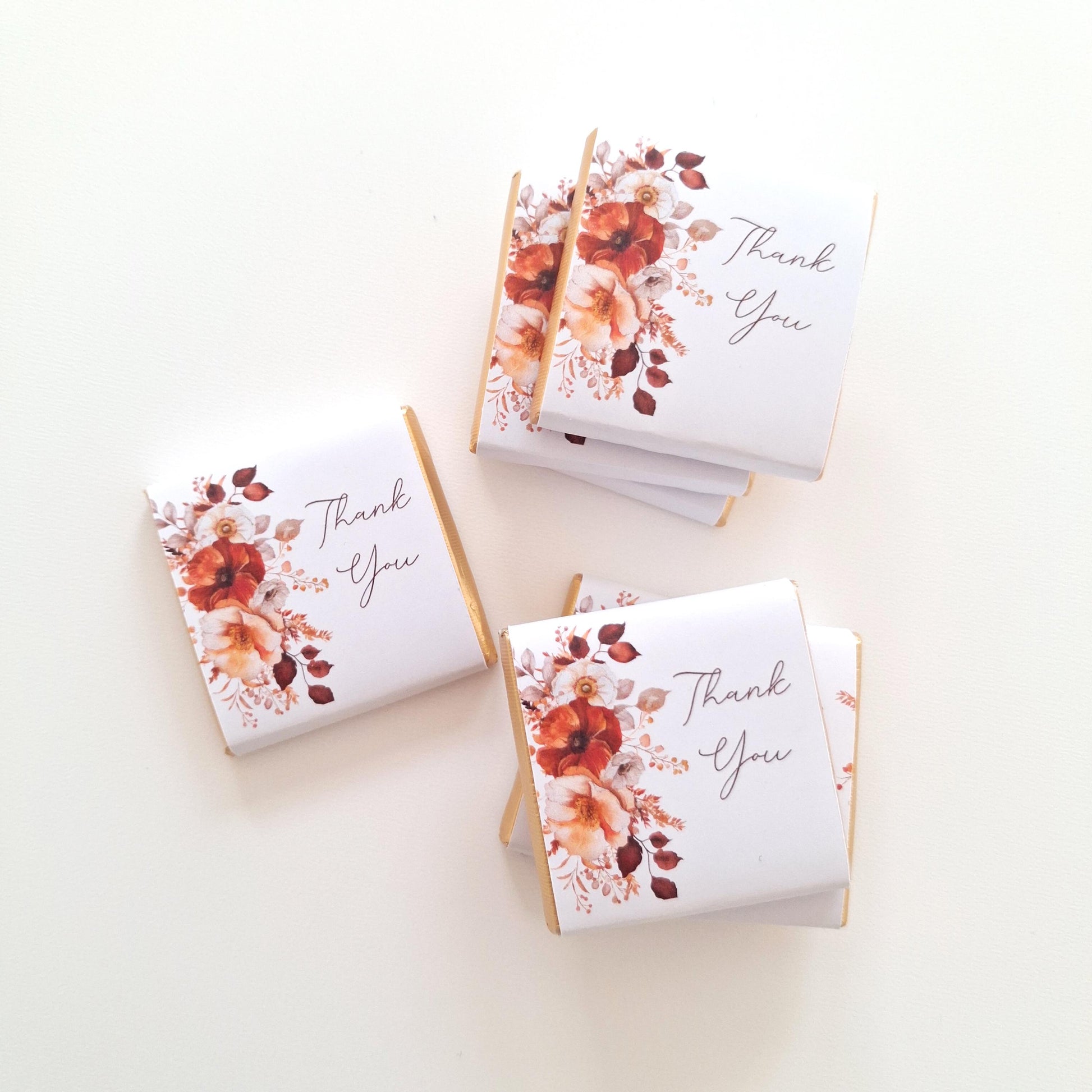 May include: Several small, white chocolate bars with gold edges are arranged on a white surface. Each bar is wrapped in white paper with a floral design in shades of red, orange, and cream, and the words "Thank You".