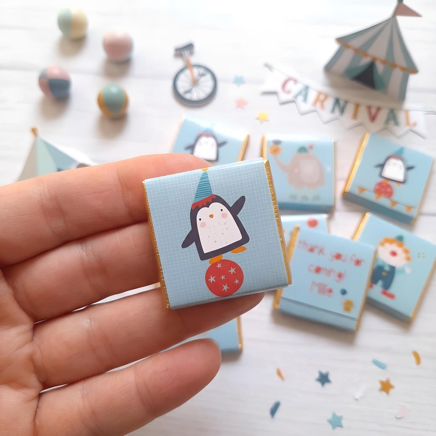 May include: A light blue square candy wrapper with a cartoon penguin balancing on a red ball. The wrapper has a gold border and is part of a set of circus-themed treats. Other wrappers feature circus animal designs.