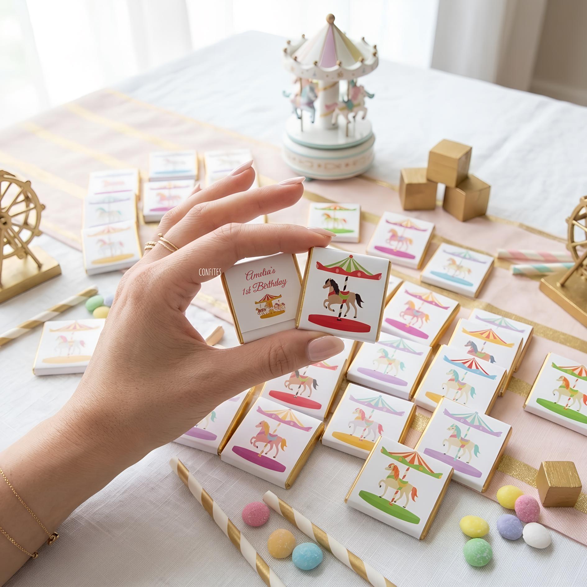 May include: A selection of square chocolates with carousel horse designs and gold edges. One chocolate is held, displaying the text "Amelia's 1st Birthday". Other chocolates are arranged on a pink and white striped surface, with a carousel music box and gold decorations.